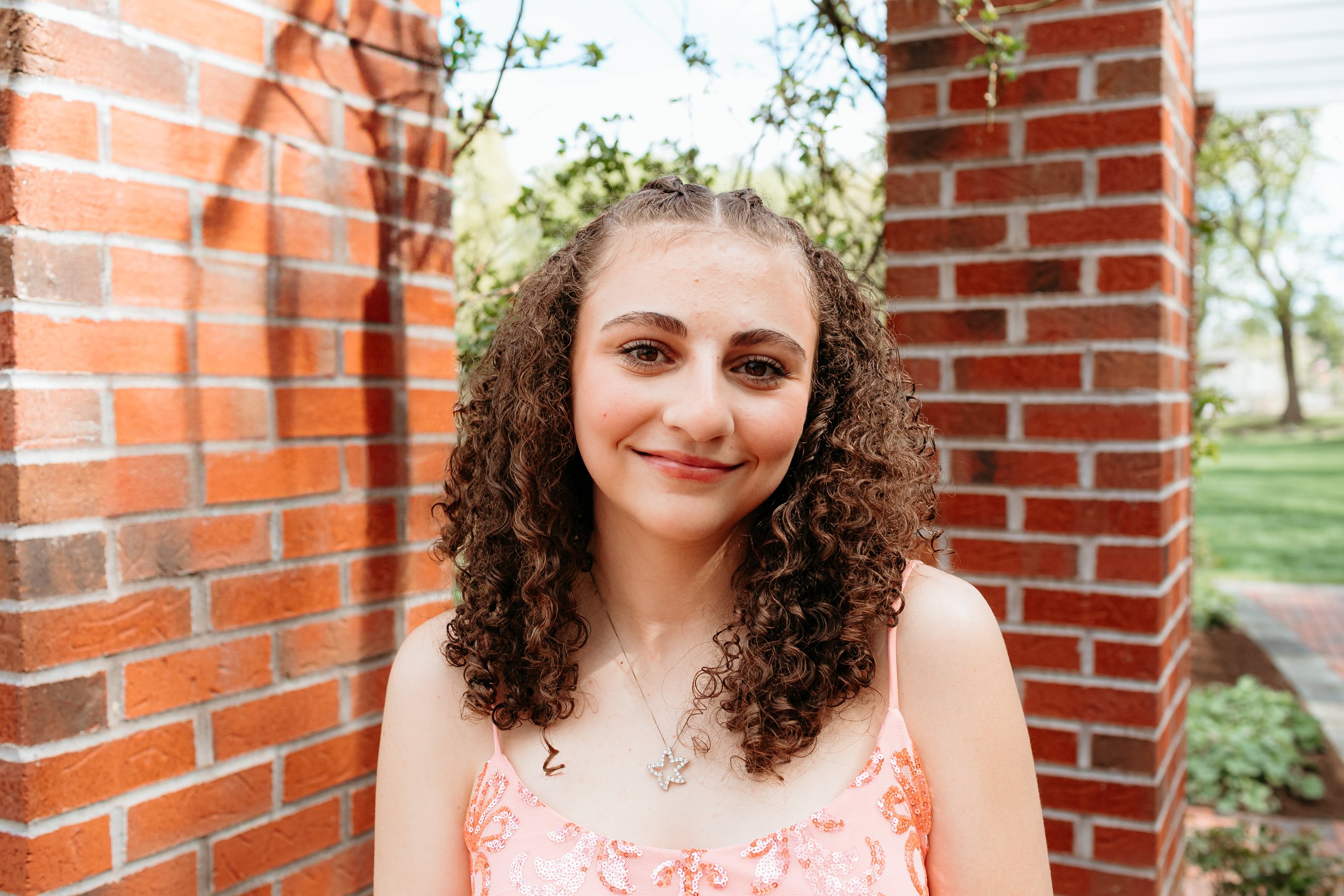 Young woman with curly hair in a pink dress, standing outdoors next to a brick wall and greenery.