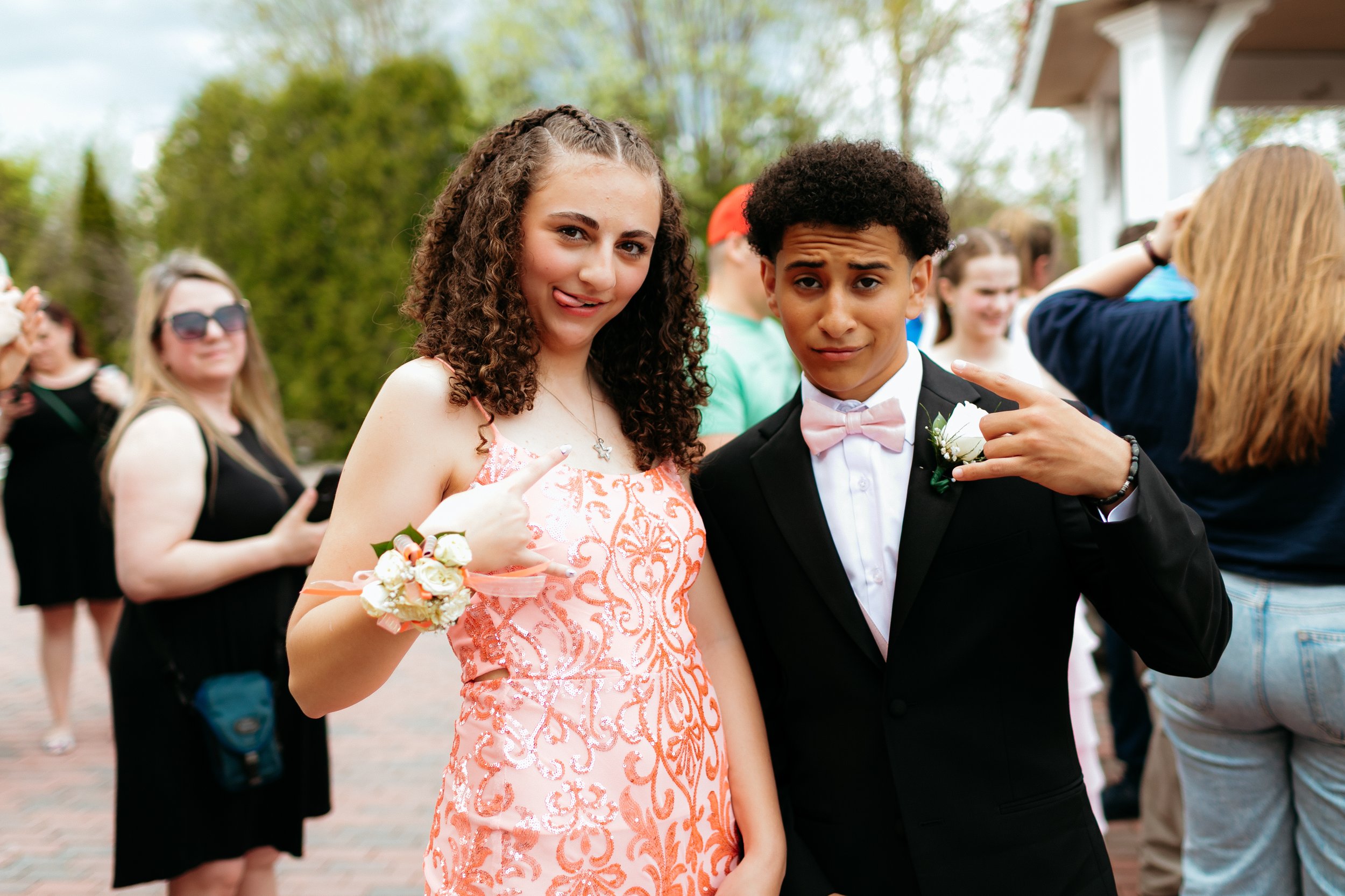 A young woman in an embellished coral dress and a young man in a black suit with a pink bow tie are posing playfully. Both are wearing corsages. People are socializing in the background outside.