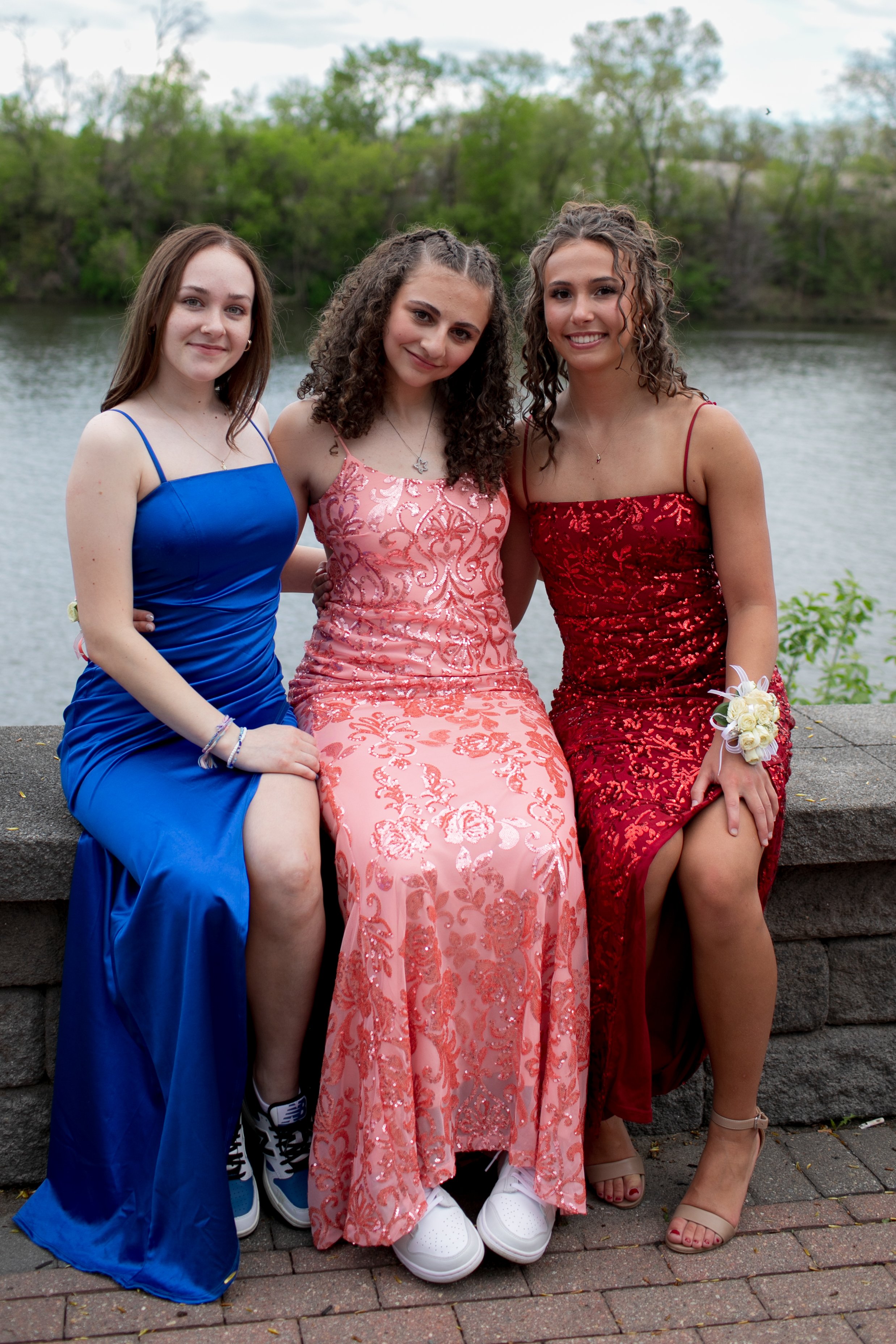 Three young women in formal gowns sitting on a stone wall by a river, wearing blue, pink, and red dresses, smiling and posing for a photo.