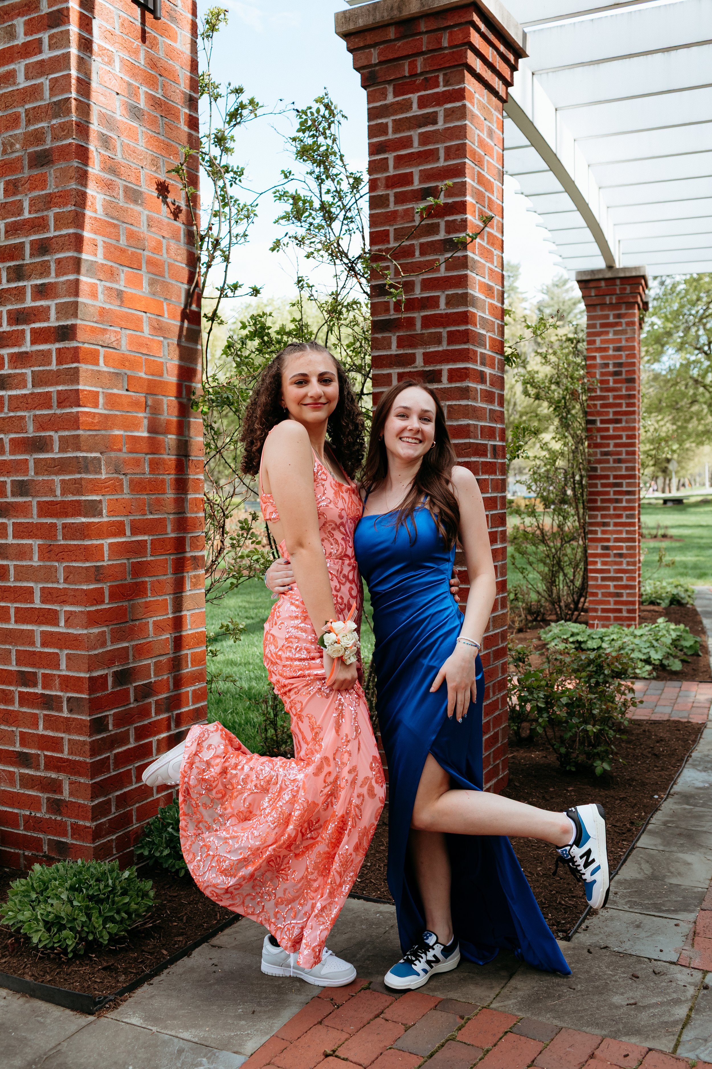 Two women in formal dresses and sneakers posing playfully next to red brick pillars in a garden setting.