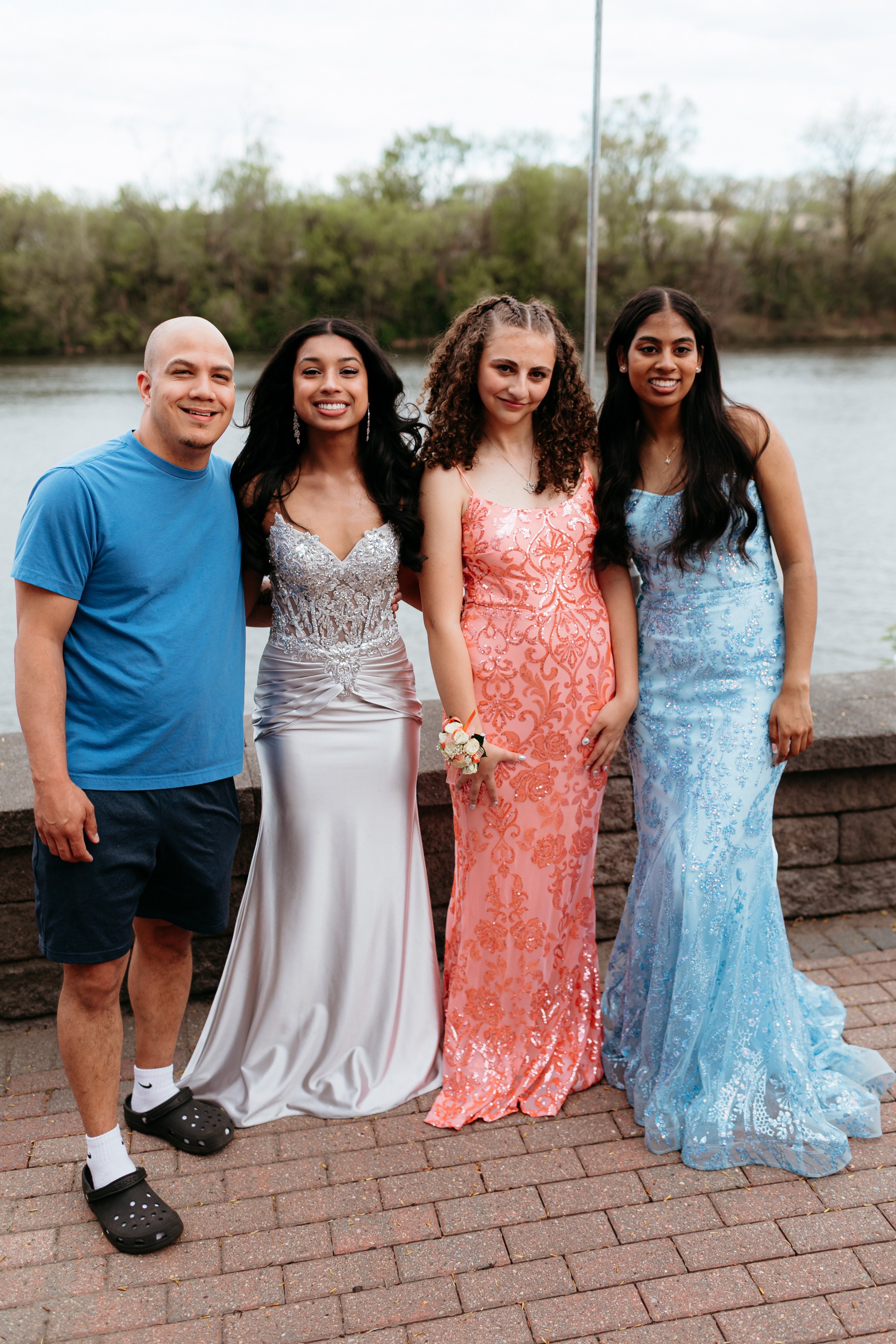 A group of three young women in formal gowns and one man in casual attire posing outdoors near a body of water.