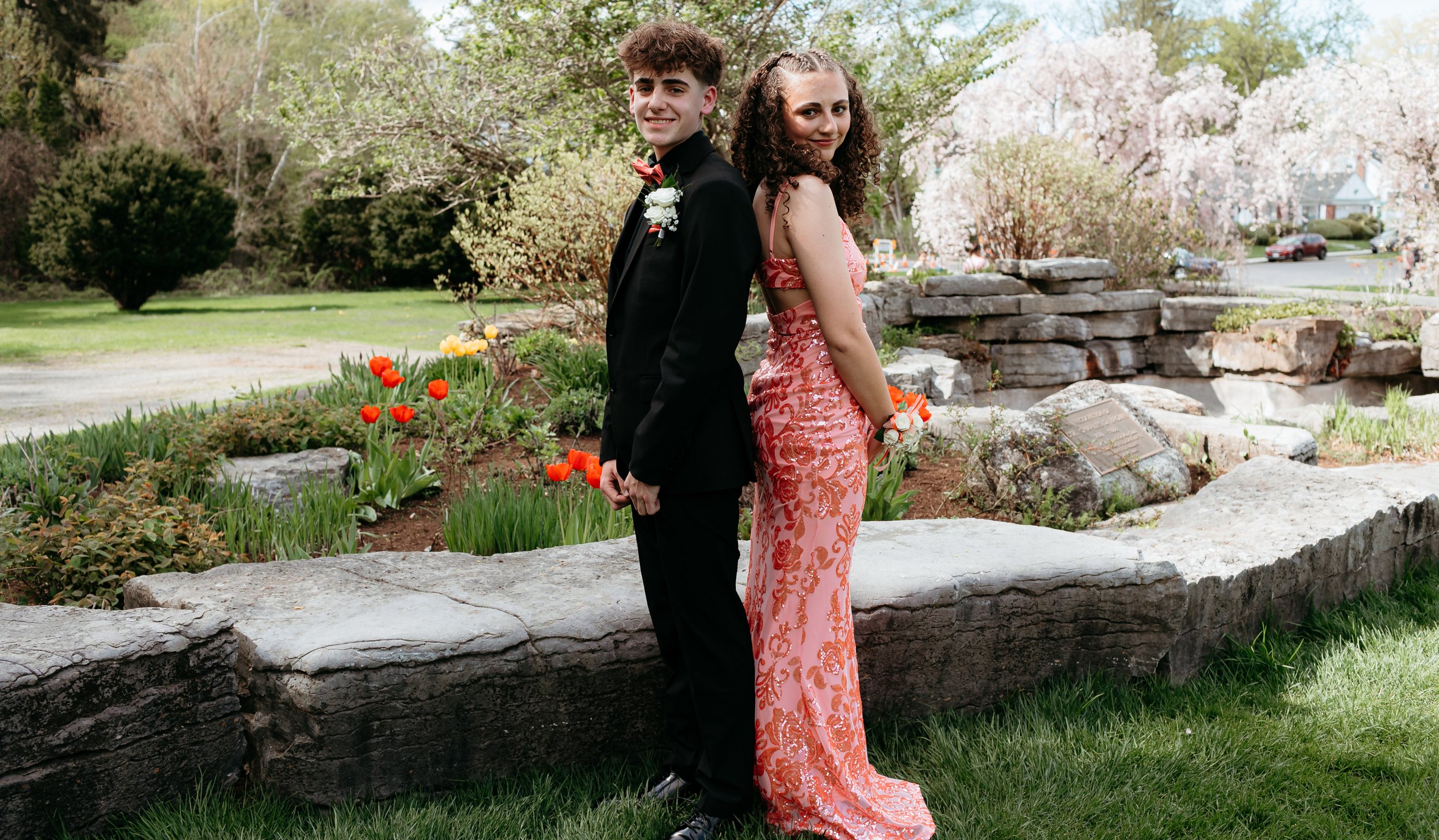 Two individuals in formal attire standing back-to-back in a garden with colorful flowers and stone landscape features.