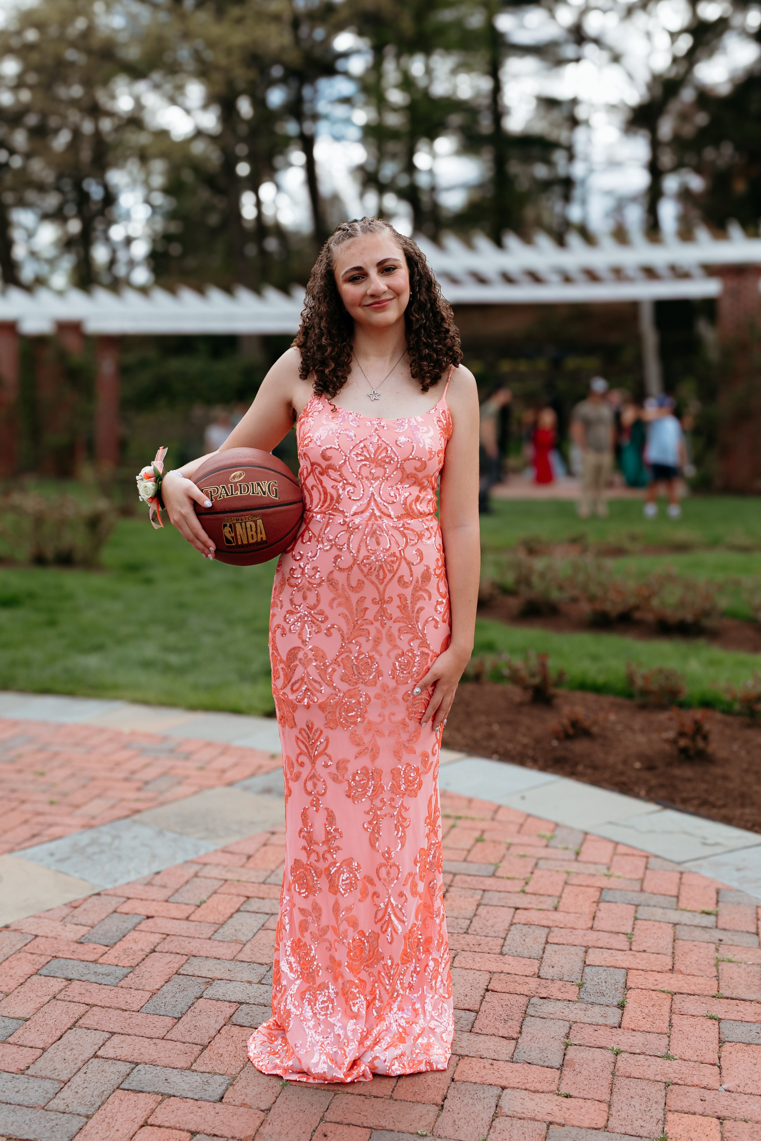 Young woman in a pink sequined gown holding a basketball outdoors on a brick pathway, with a corsage on her wrist, greenery and trees in the background.