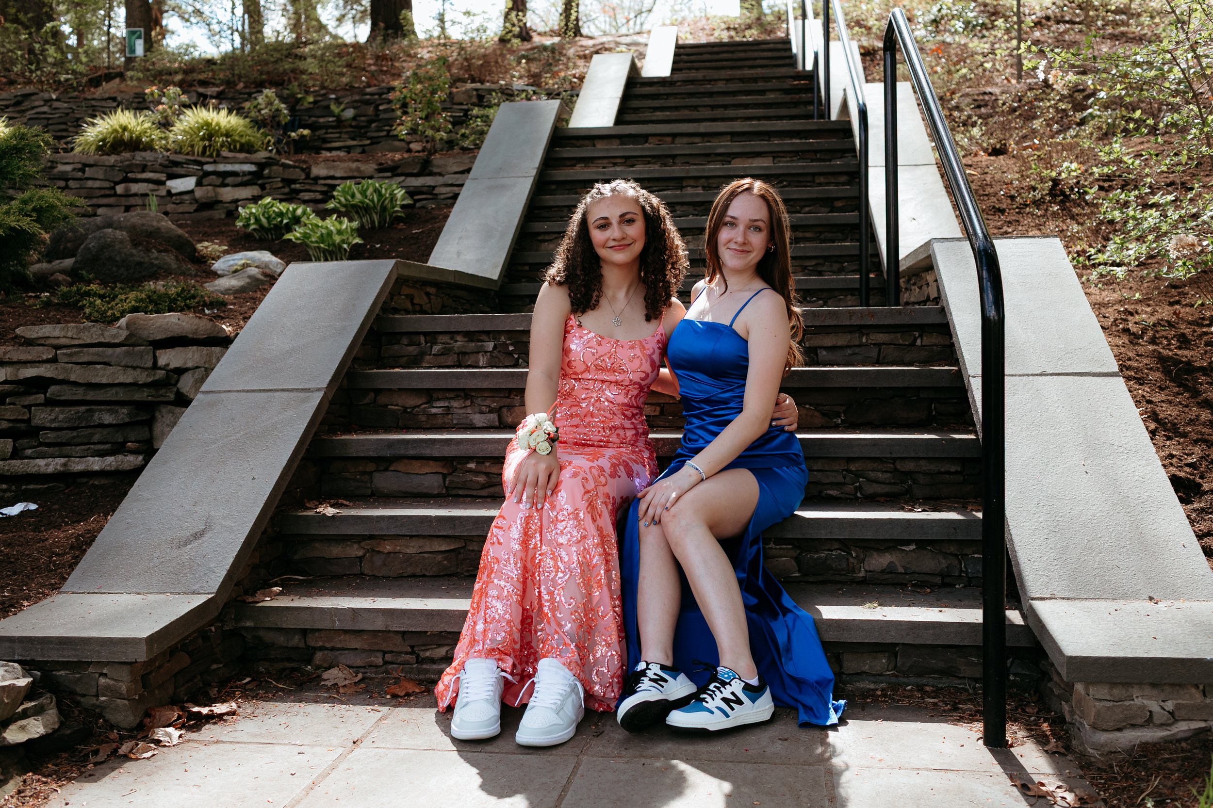 Two young women in formal dresses sitting on outdoor stone stairs, with one in a pink dress and the other in a blue dress, both wearing sneakers. They are in a garden setting with stone wall and plants around.