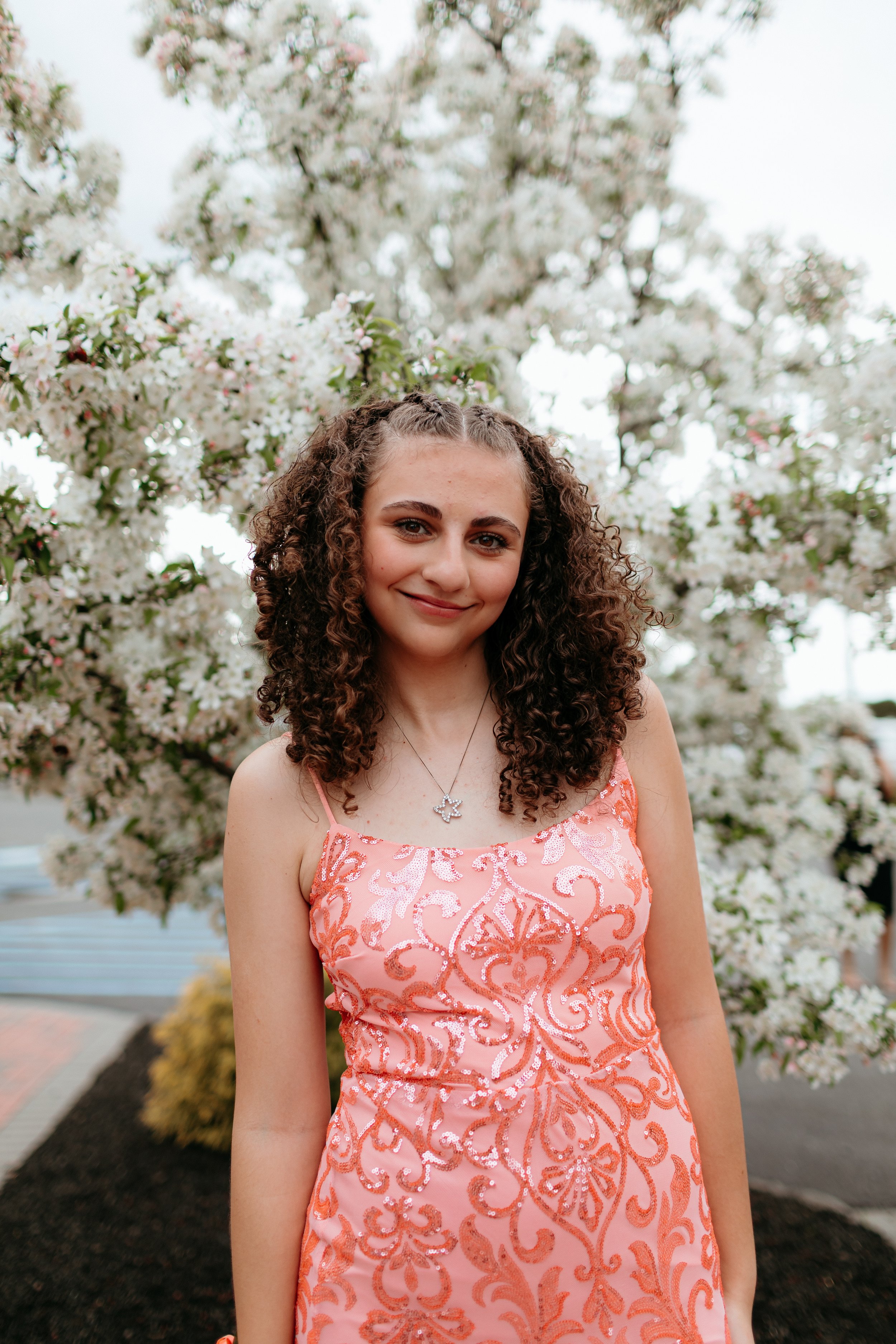 Young woman in pink floral dress smiling in front of flowering tree.