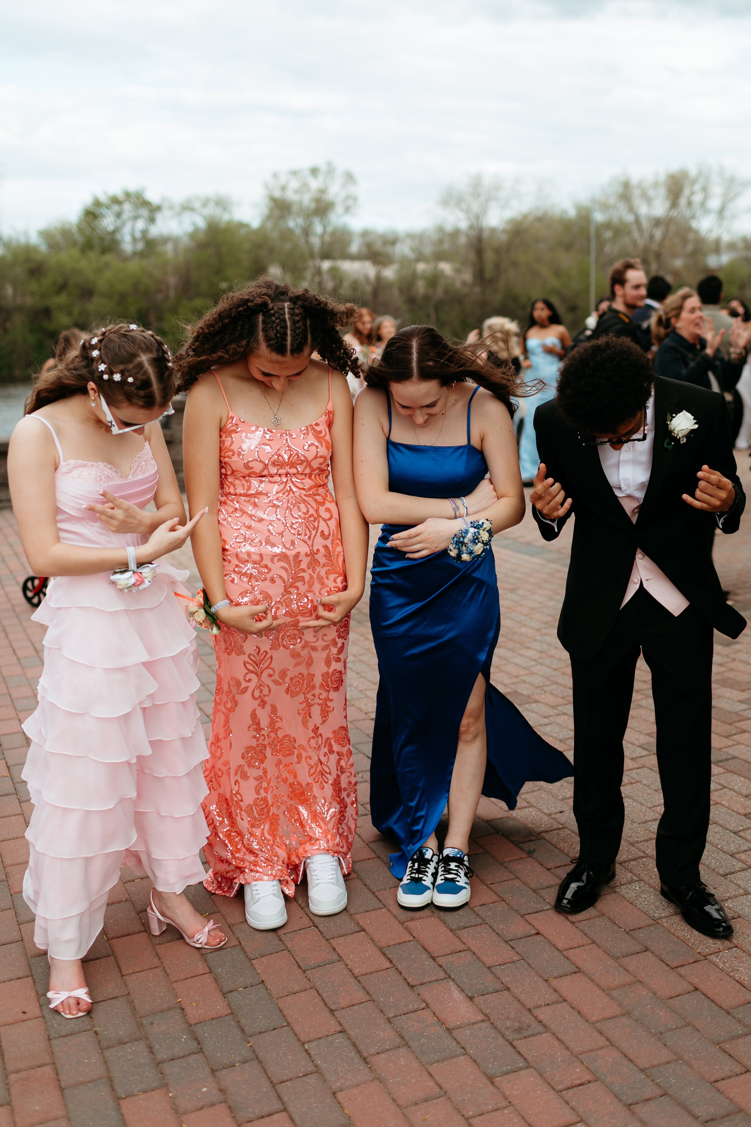 Group of teenagers in prom attire looking down at their shoes, standing on a brick pathway.