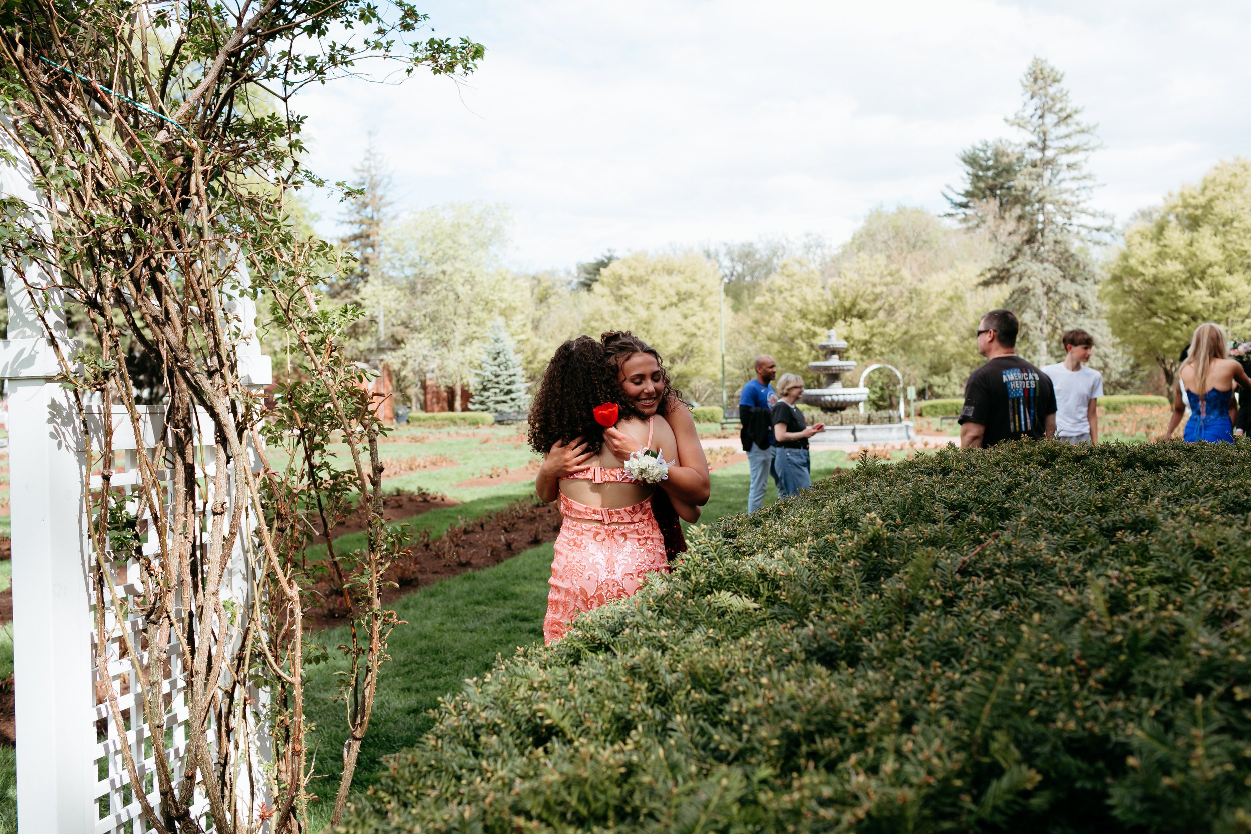 Two people hugging in a garden with a fountain in the background and others walking around.