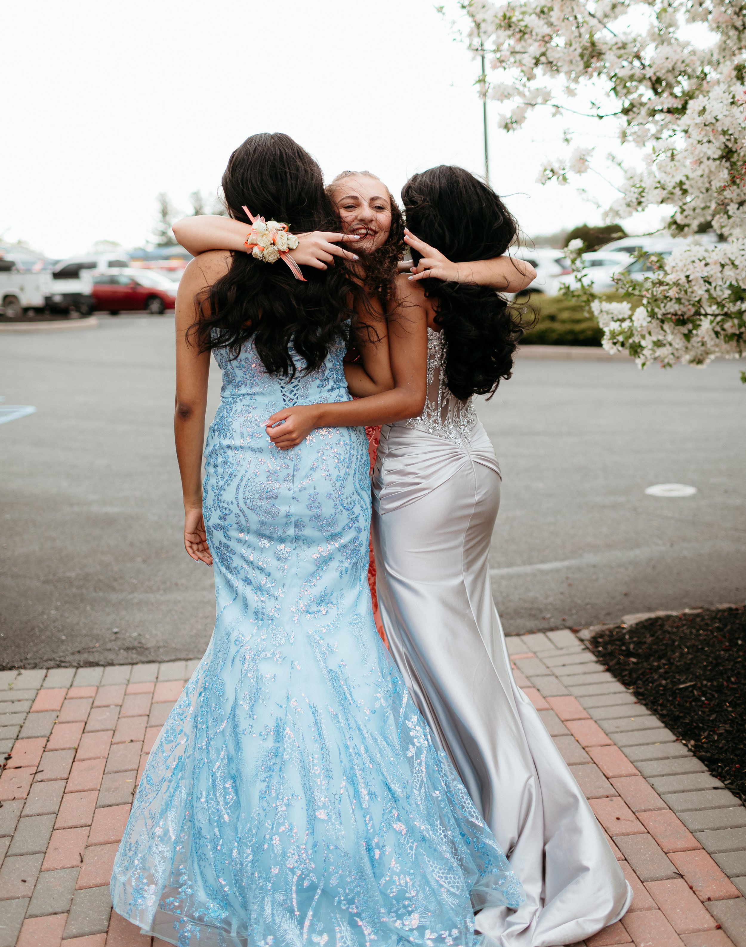 Three young women in formal dresses hugging and posing for a photo outdoors, with one wearing a light blue gown and another in a silver gown. A flowering tree is visible in the background.