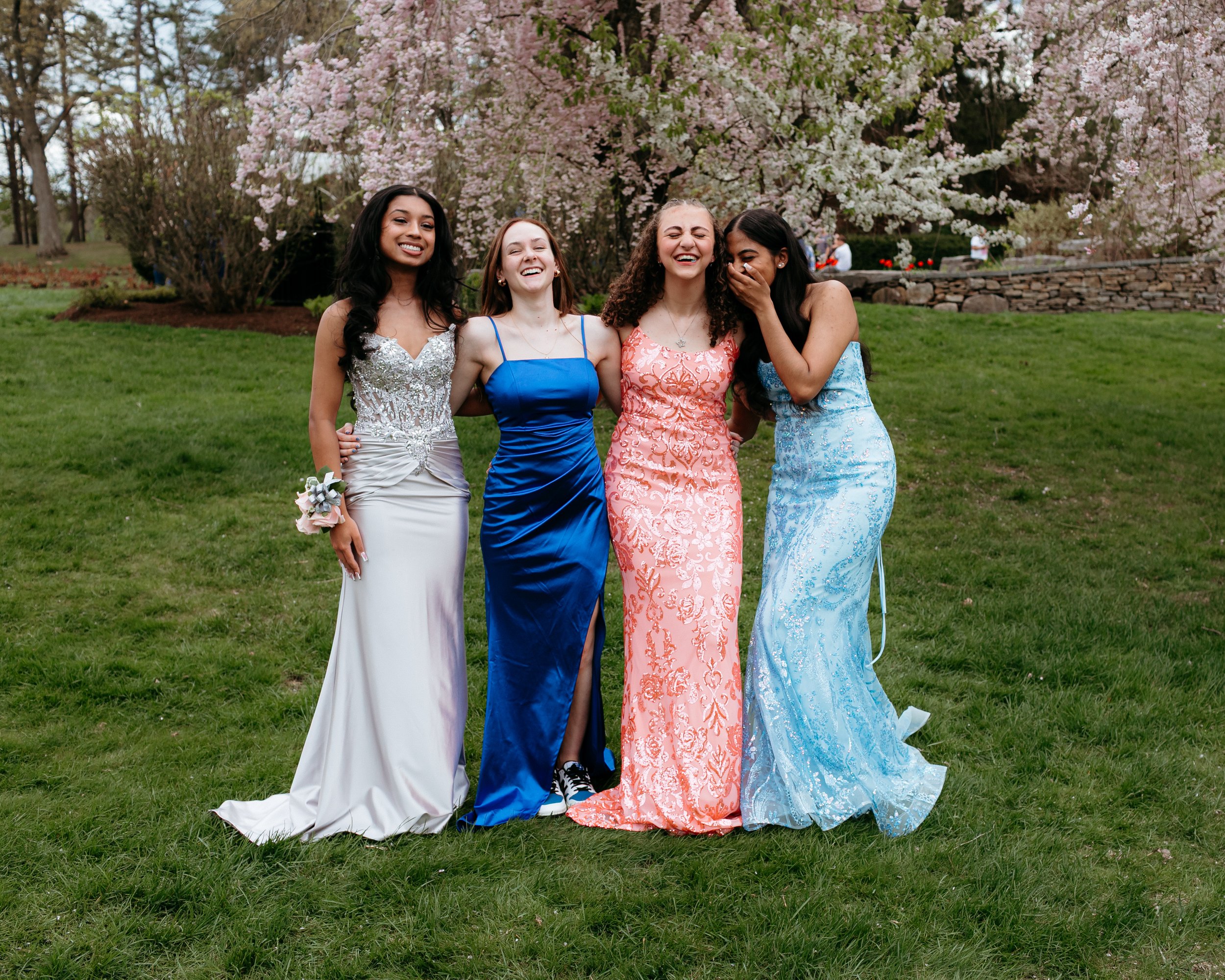 Four young women in formal dresses standing together on grass in front of blooming trees.
