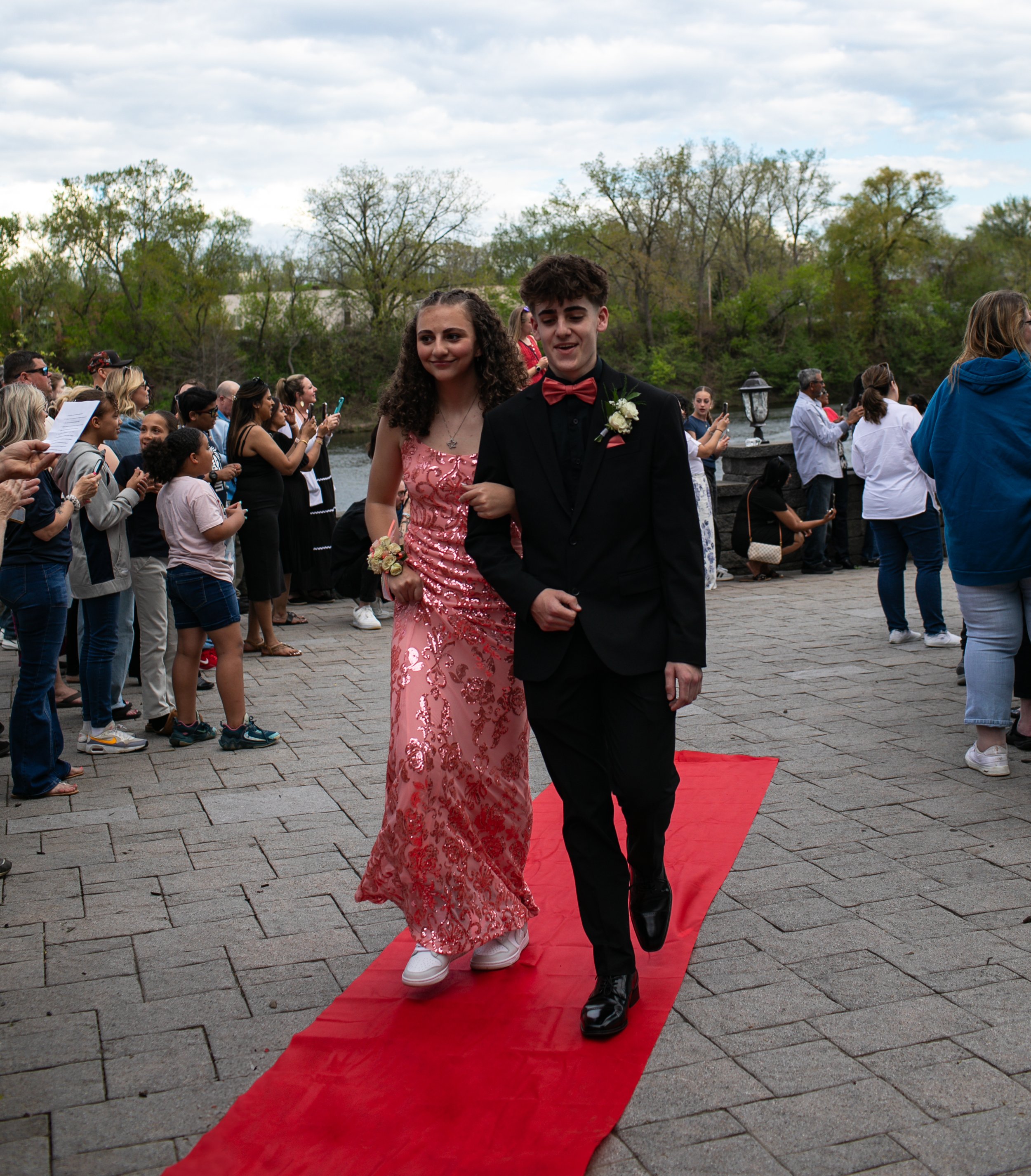 A young couple walking on a red carpet outdoors, dressed in formal attire. The girl is wearing a pink sequin dress and the boy is in a black suit with a red bow tie. A crowd is gathered around taking pictures.