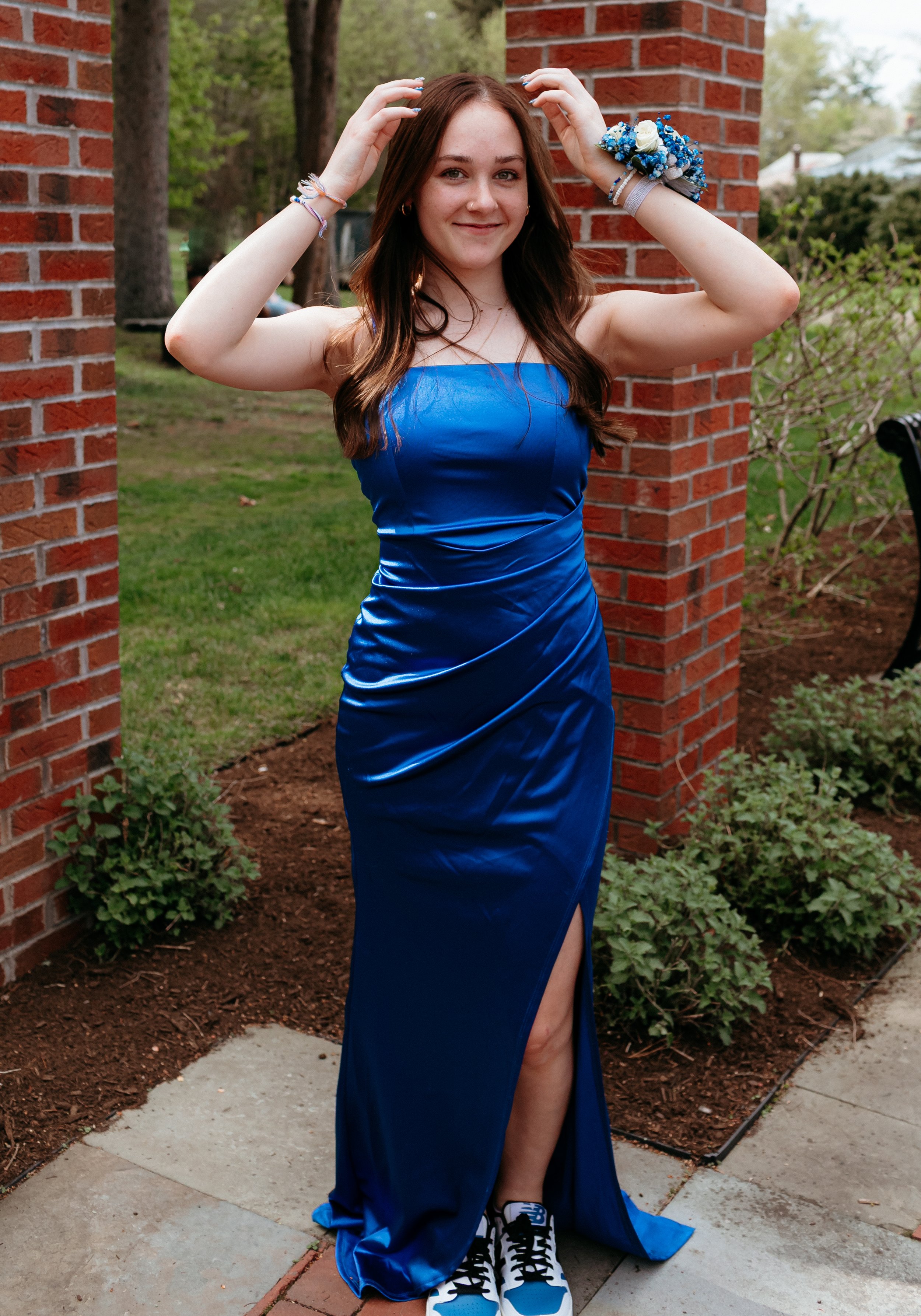 Young woman in a blue dress posing outdoors with brick columns and greenery.