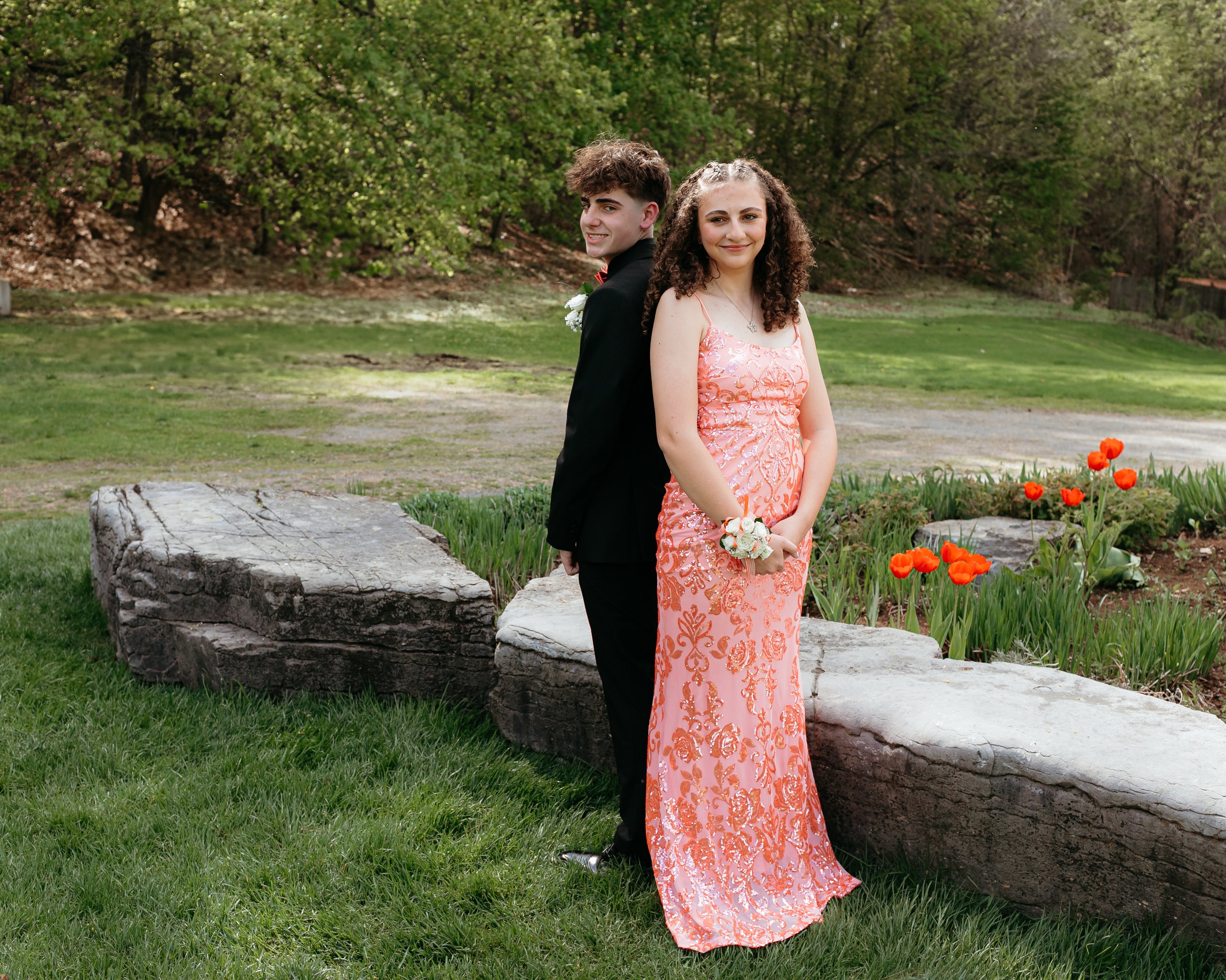 Teen couple dressed in formal attire for prom in a garden setting with stone benches and red flowers.