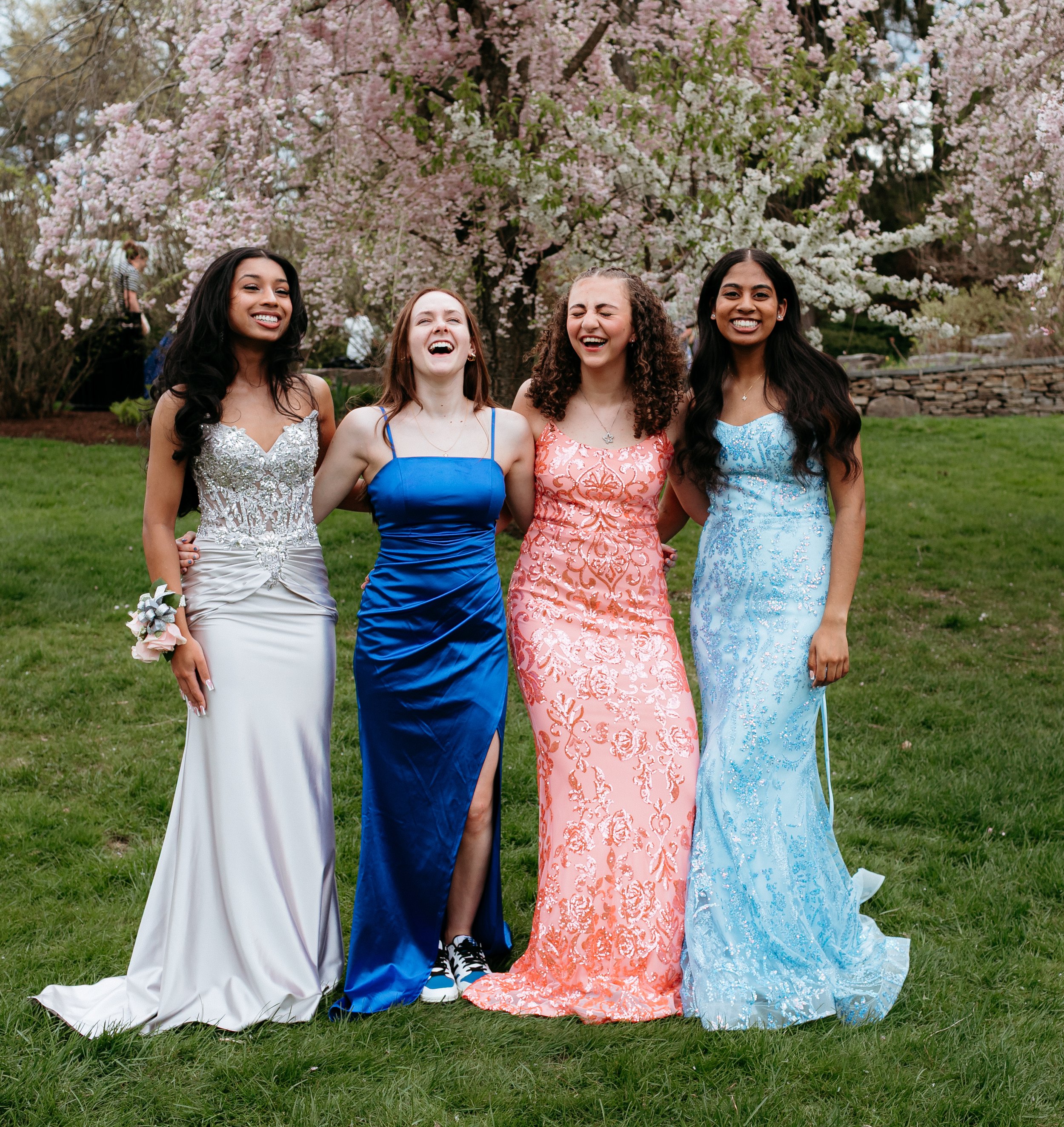 Four young women in colorful, elegant prom dresses standing on grass in front of blooming trees, smiling and holding each other.