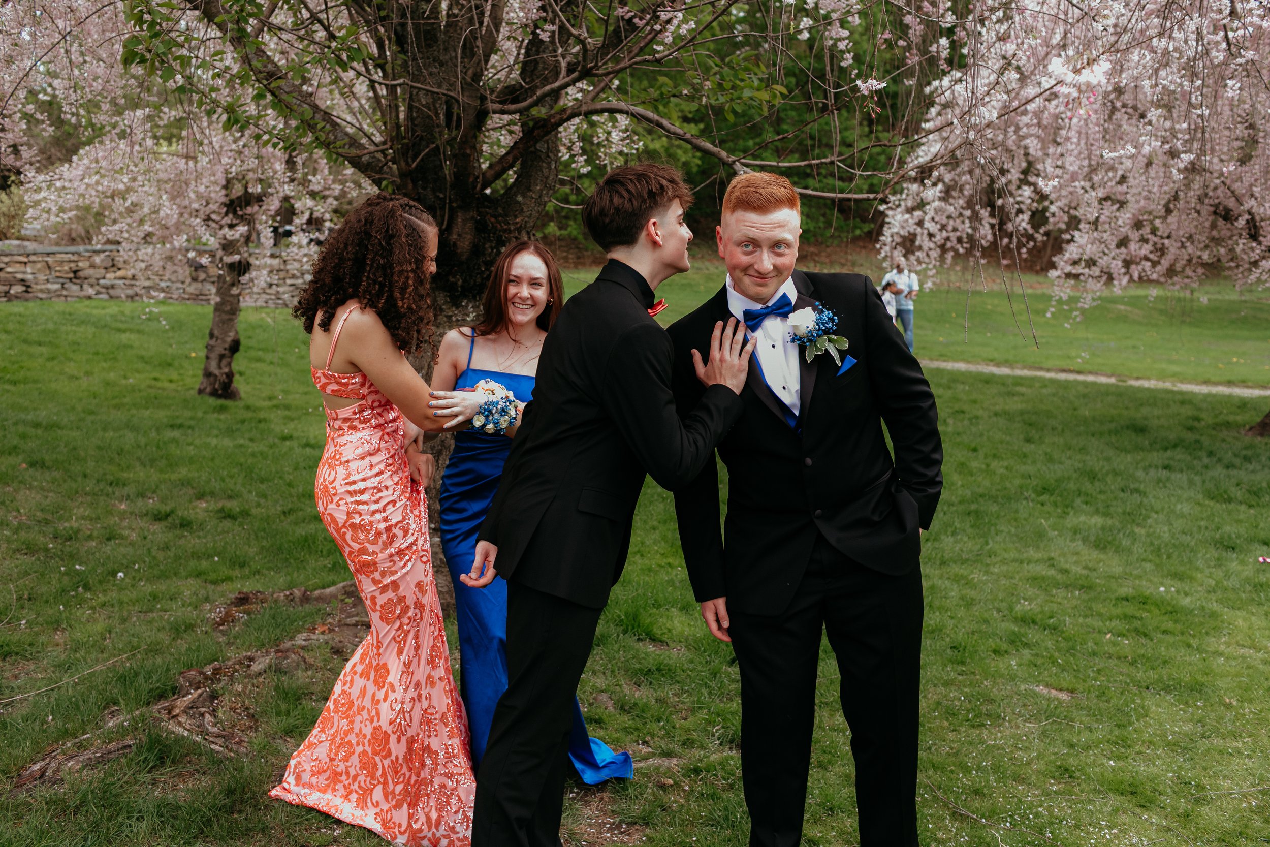 Group of young people in formal attire posing playfully outdoors with flowering trees in the background.