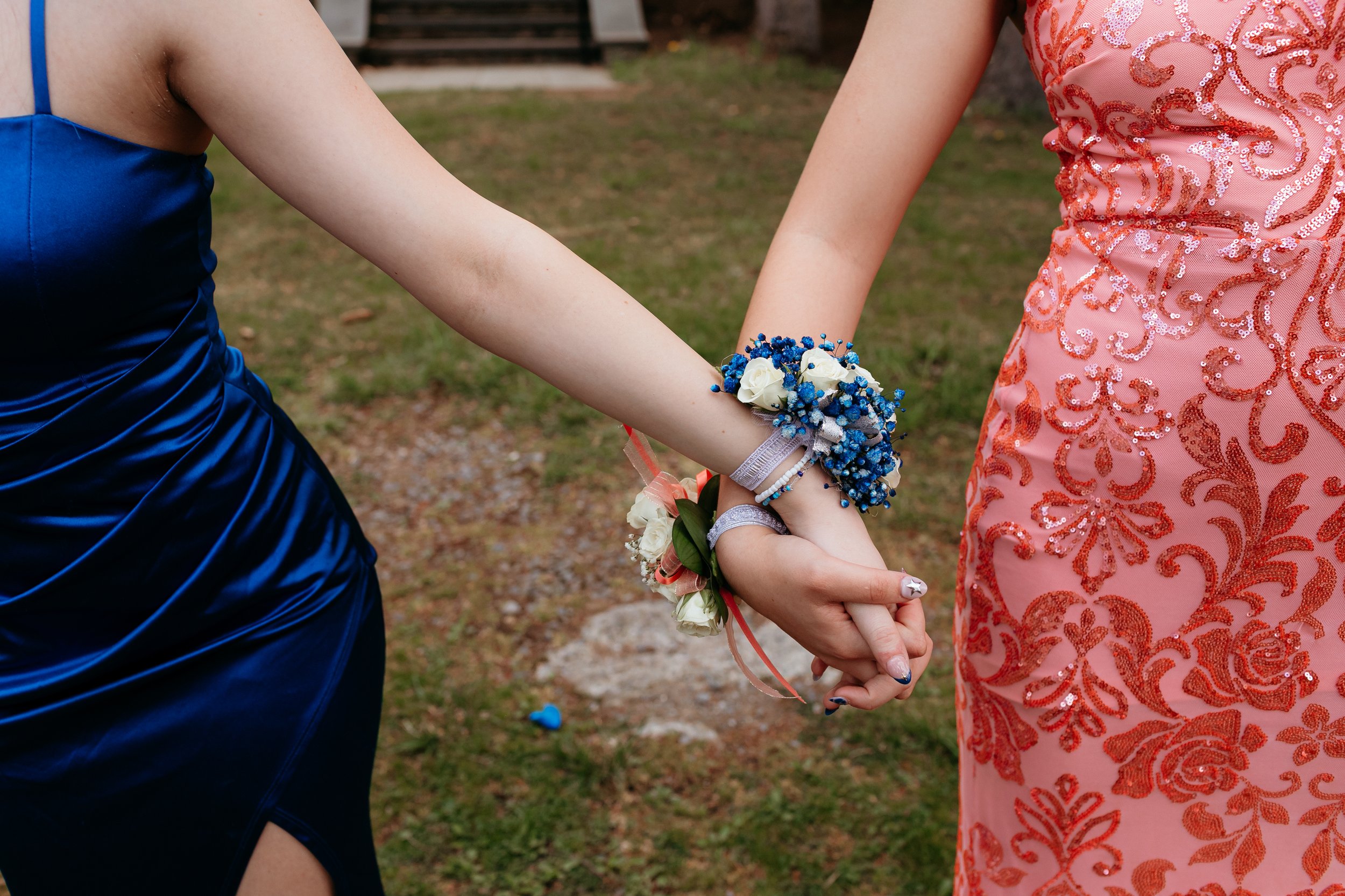 Two people in formal dresses holding hands, wearing corsages. One dress is blue, the other is pink with sequin patterns.