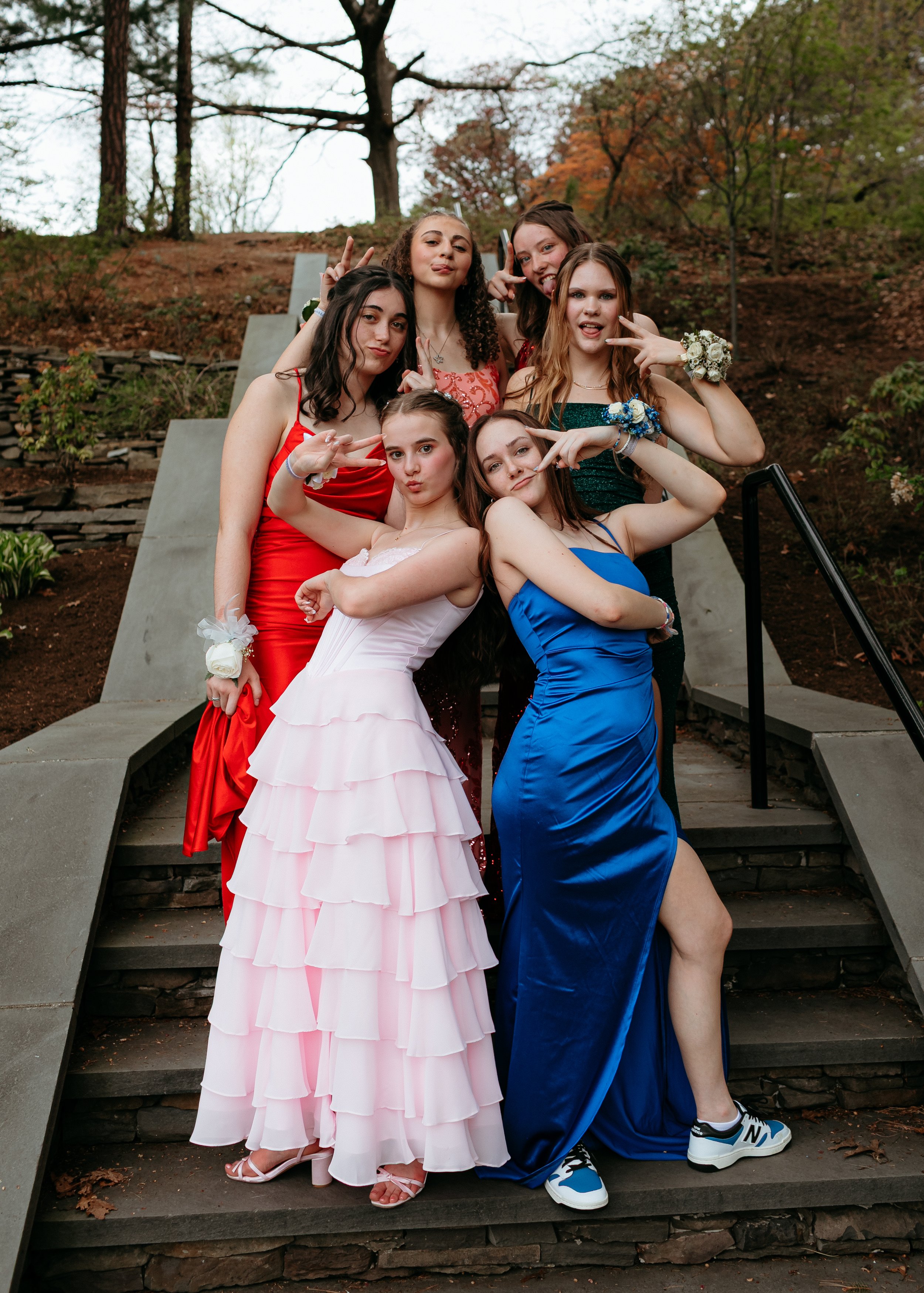 A group of six young women posing on outdoor stairs, wearing vibrant formal dresses in various colors. They are striking playful poses and making peace signs with their hands. The background includes trees and a rustic stone wall.