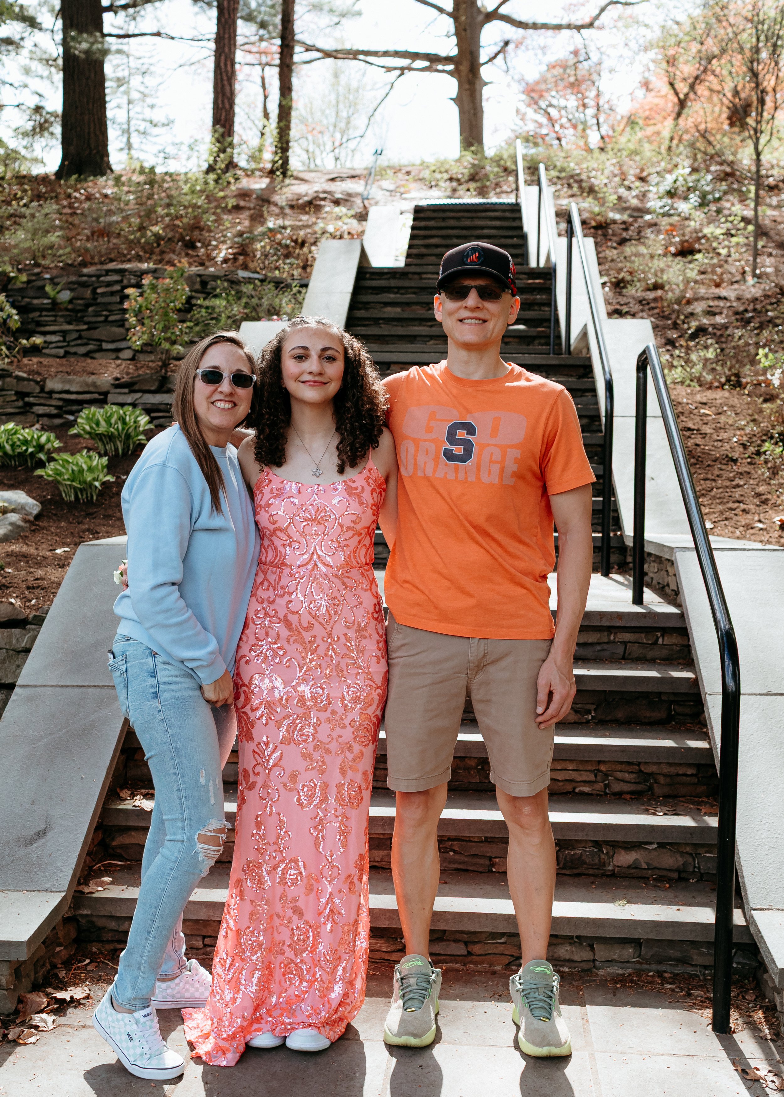Three people standing on outdoor steps with trees and plants in the background. The person in the center is wearing a pink dress, the person on the left is in a blue jacket and jeans, and the person on the right is wearing an orange "Go Orange" T-shi