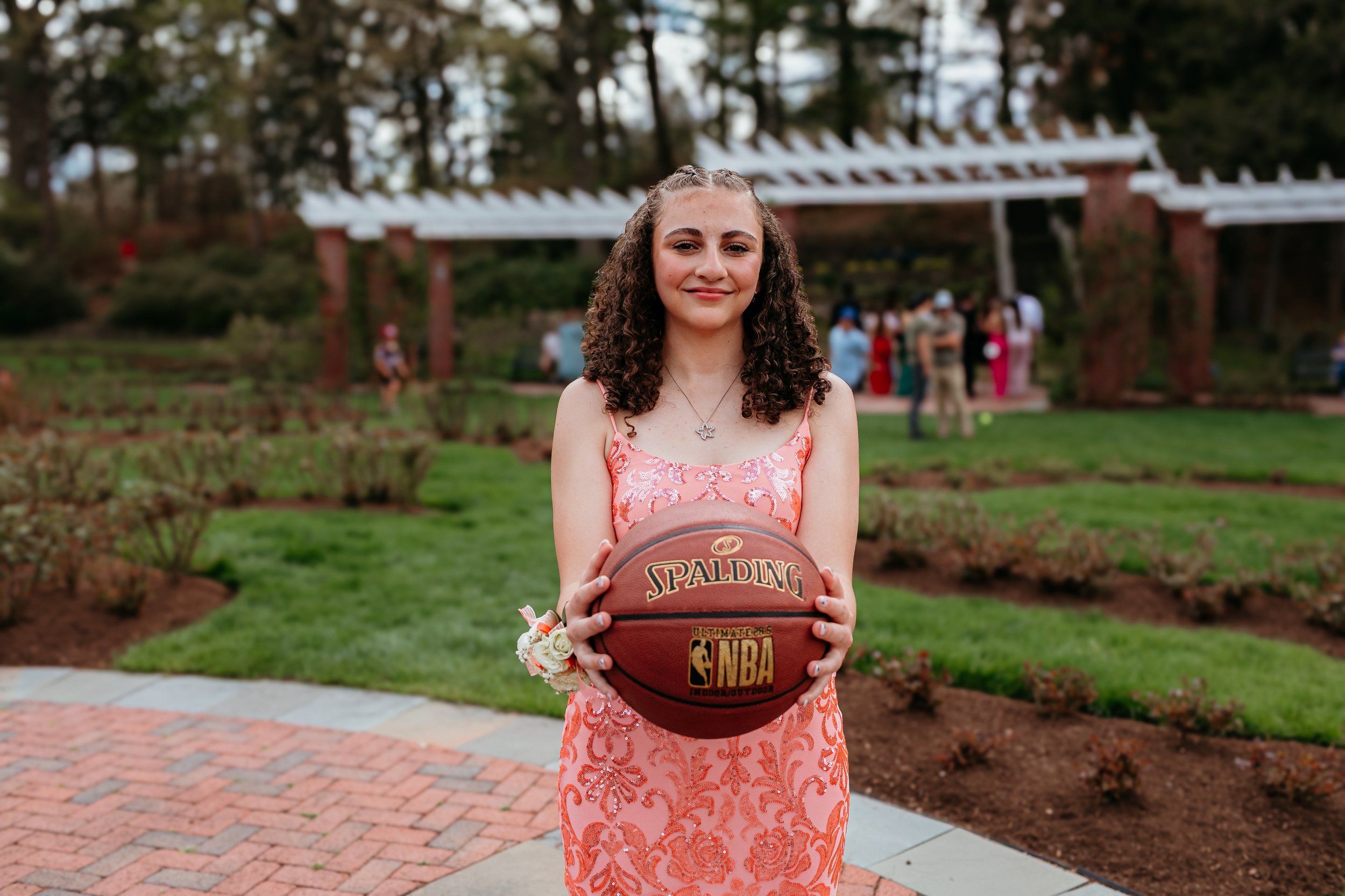 Young woman in a floral dress holding a Spalding NBA basketball in a garden setting with a white pergola in the background.