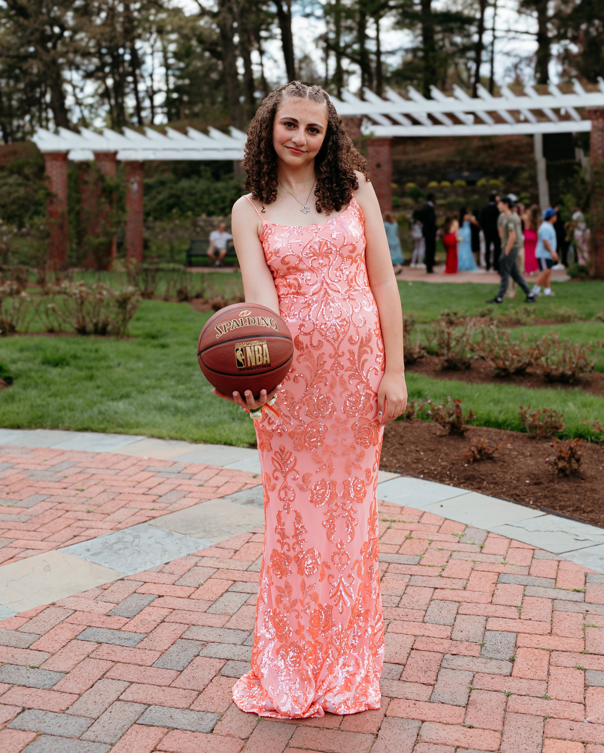 Young woman in a peach-colored formal gown holding a basketball, standing on a brick path in a garden setting.