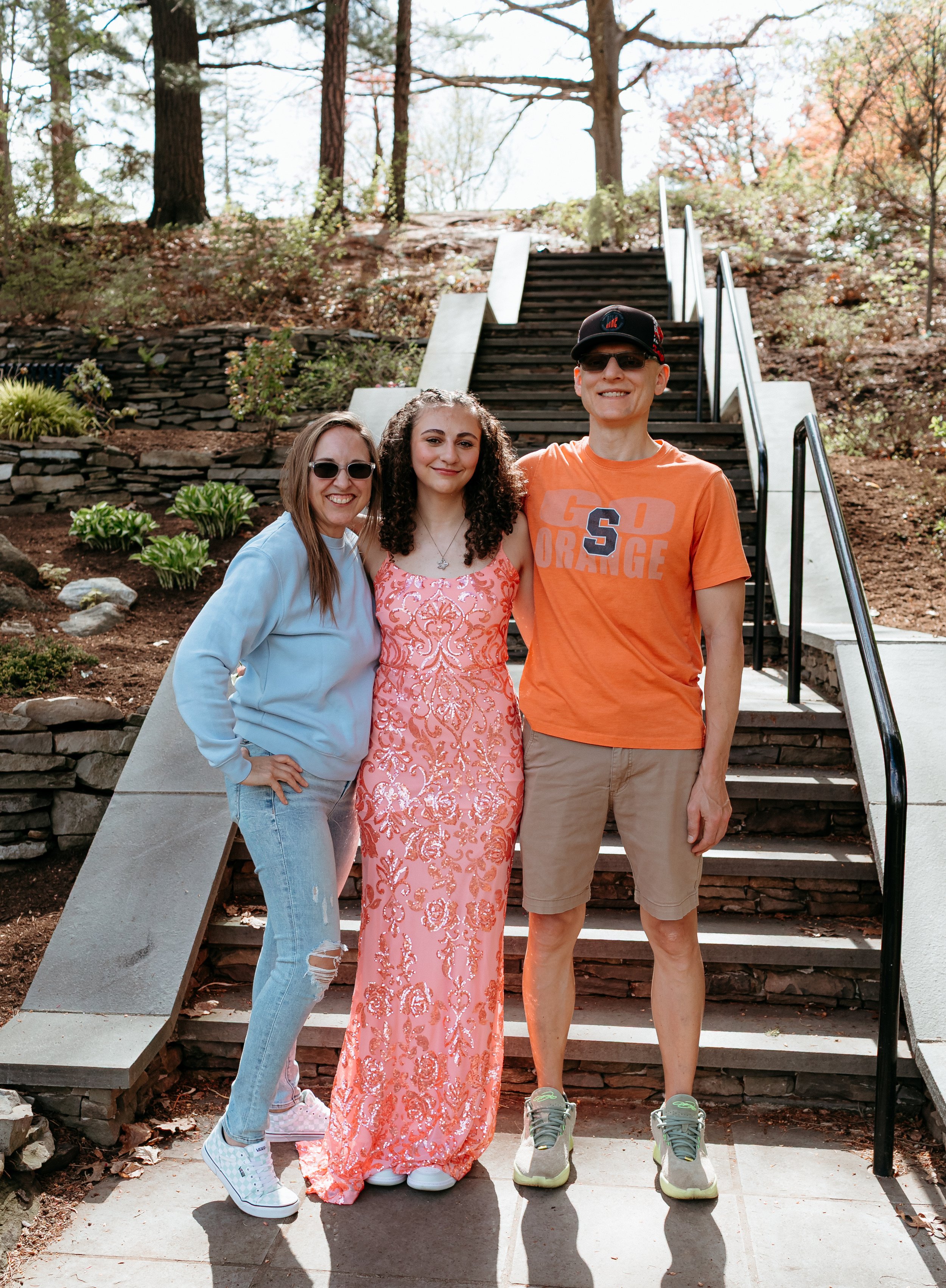 Three people posing outdoors on stone steps; one in a blue hoodie and jeans, one in a pink dress, and one in an orange t-shirt and shorts, surrounded by trees and greenery.