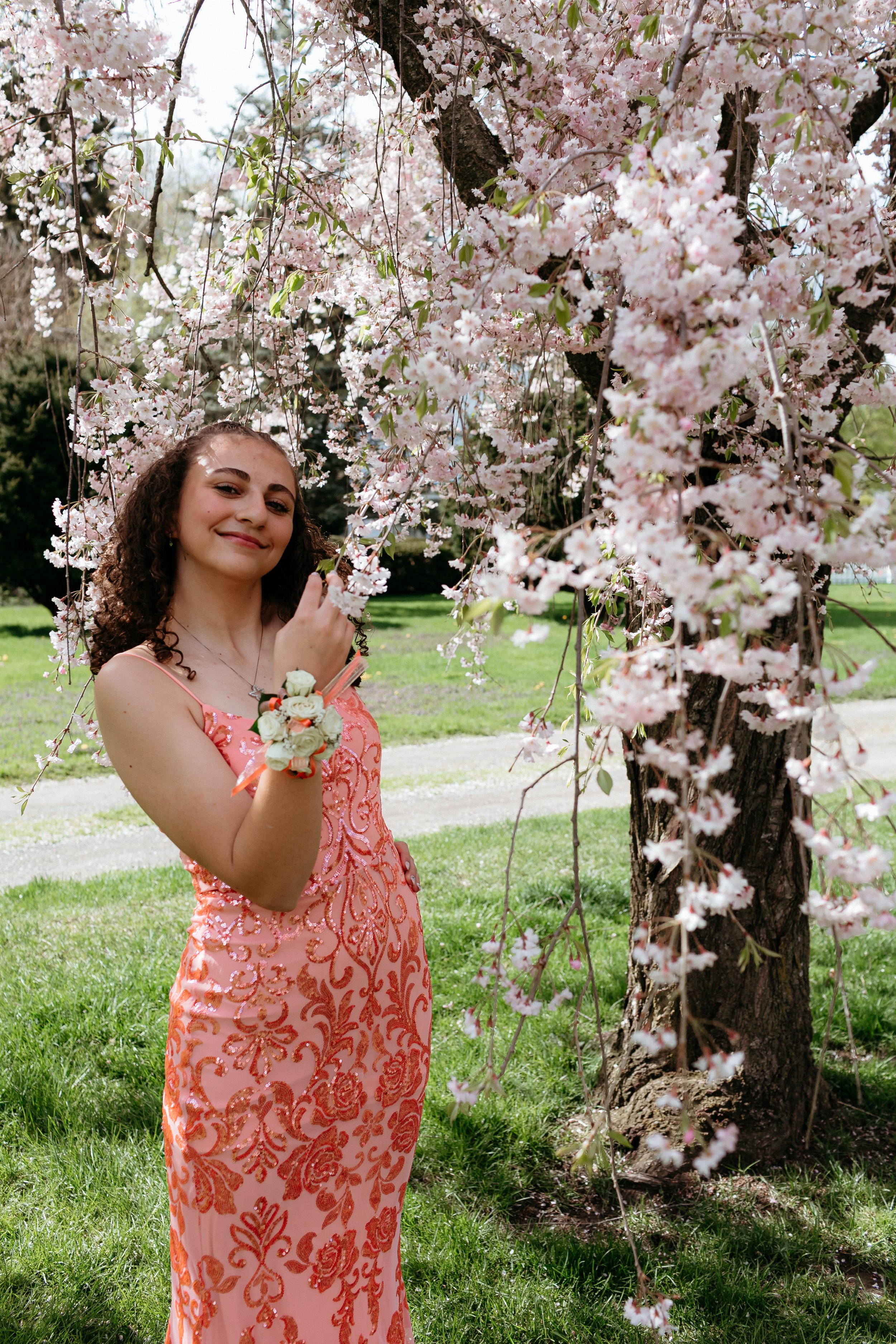 Young person in a floral pink dress standing near a blossoming cherry tree branch with a corsage on their wrist.