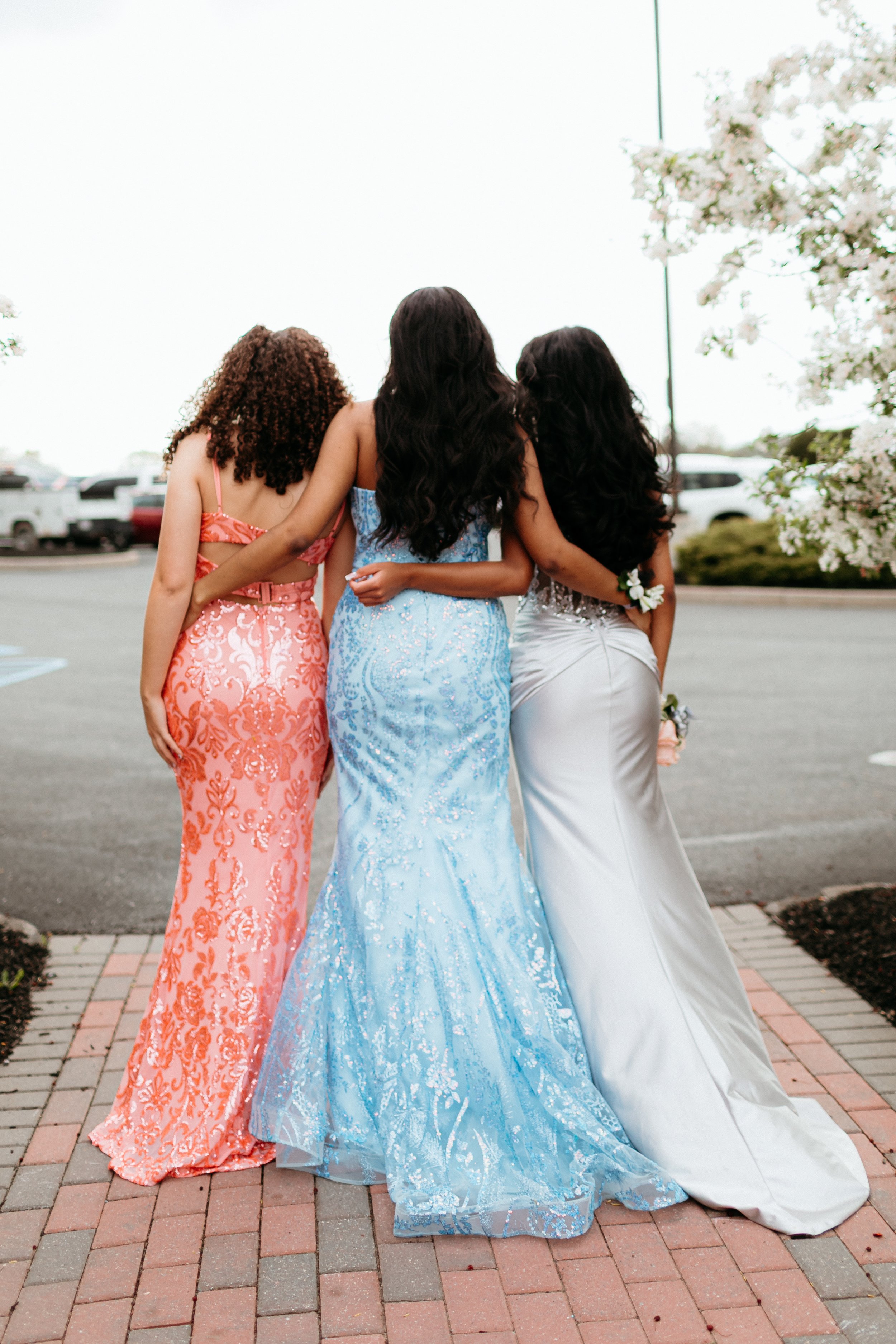 Three women in formal gowns standing with their backs to the camera, one in a coral dress, one in a blue dress, and one in a white dress, outdoors on a paved area.