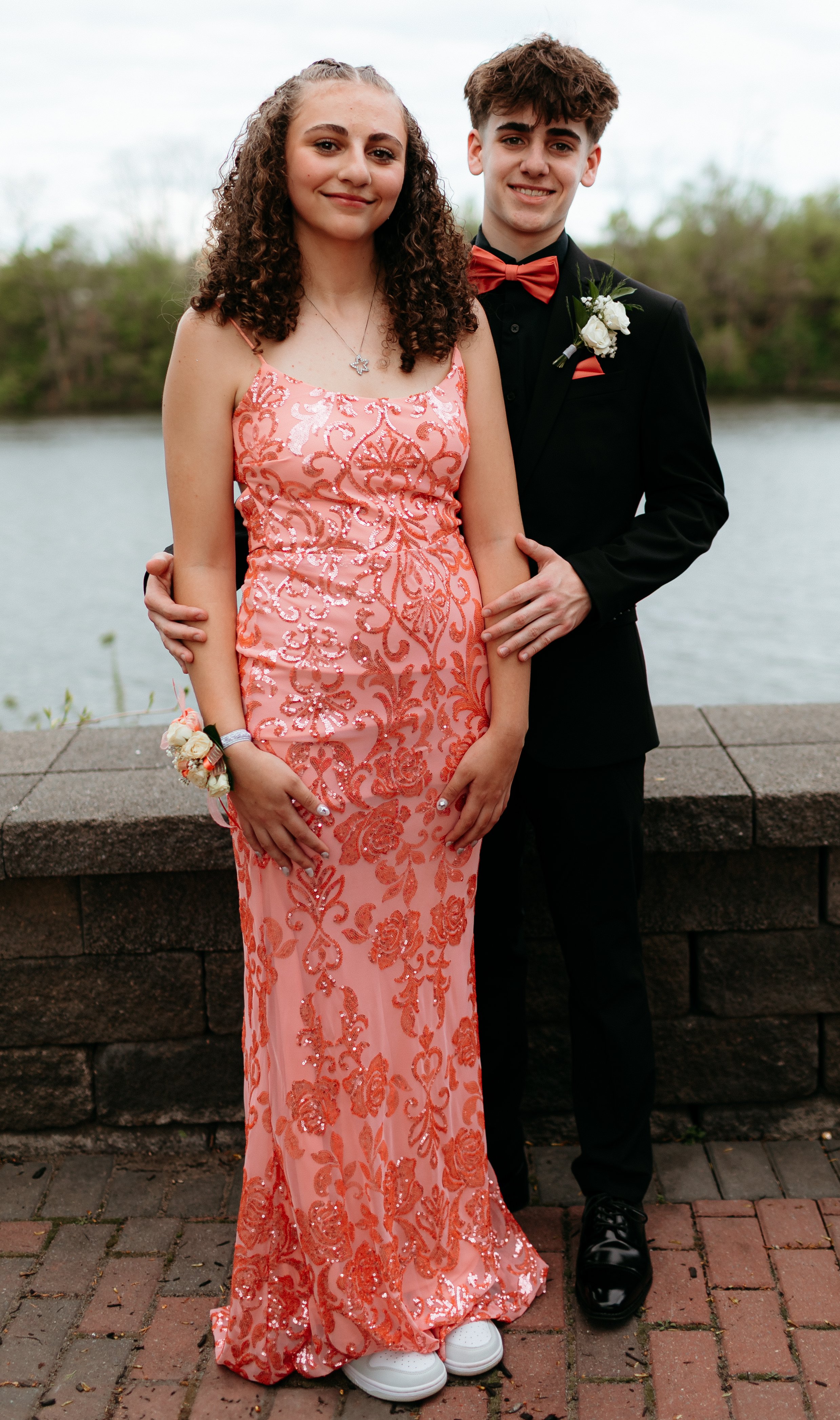 A young couple dressed for a formal event. The girl is wearing a pink sequin gown and white sneakers, with a wrist corsage. The boy is in a black suit with a red bow tie and boutonniere. They are standing by a stone wall with water in the background.