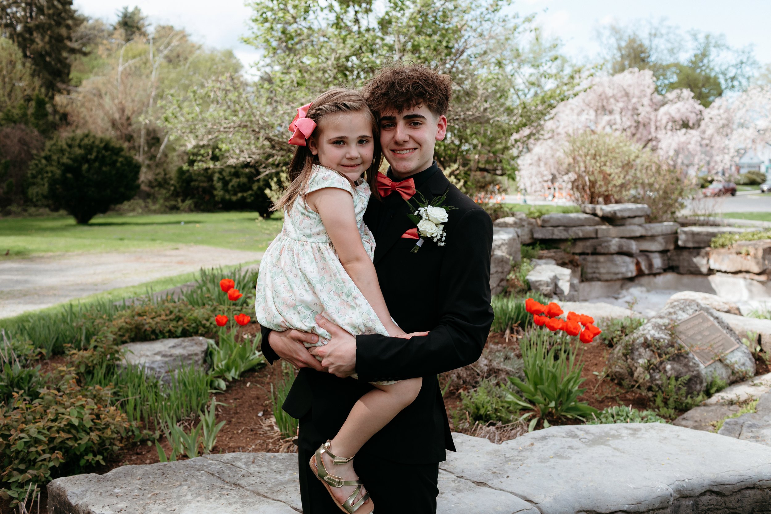 A young man in formal attire holding a young girl in a garden, surrounded by greenery and red flowers.