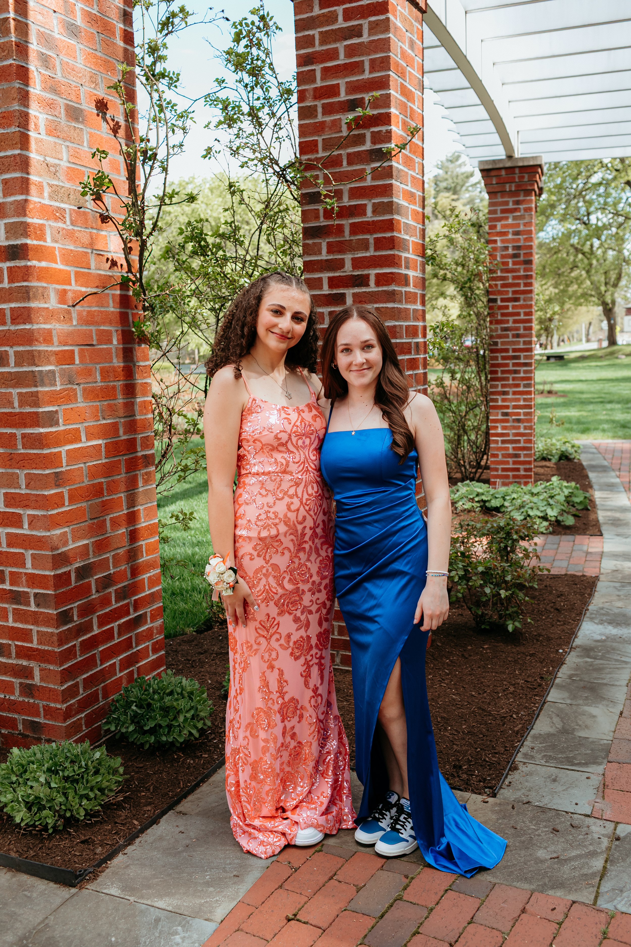 Two young women posing in formal dresses in front of a brick archway and garden. One is wearing a pink dress with floral designs and a corsage, while the other is in a blue dress with sneakers. They are standing on a brick path, surrounded by greener