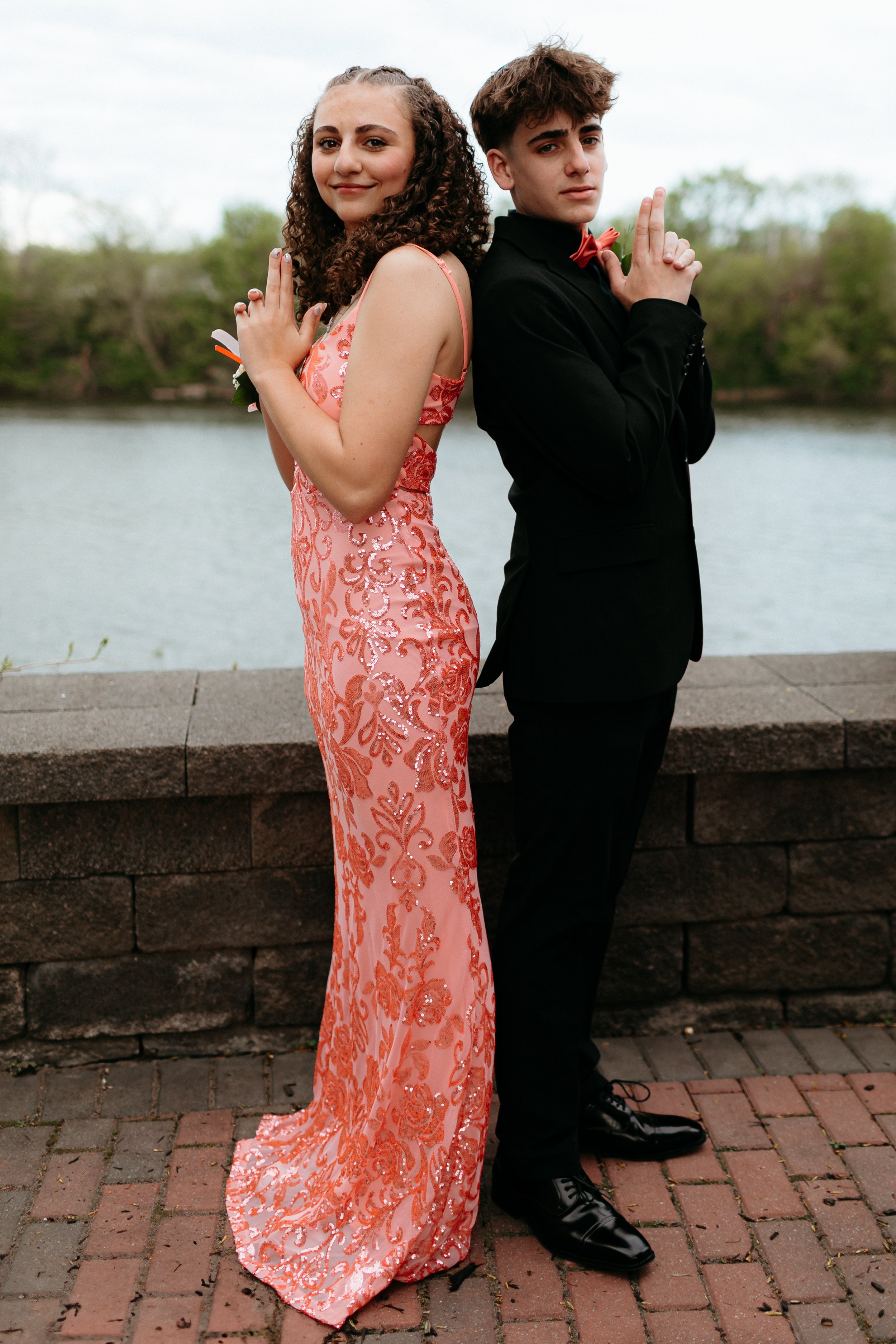 Two people posing back-to-back in formal attire near a waterfront. The woman is wearing a pink sequined dress and the man is in a black suit with a bow tie.