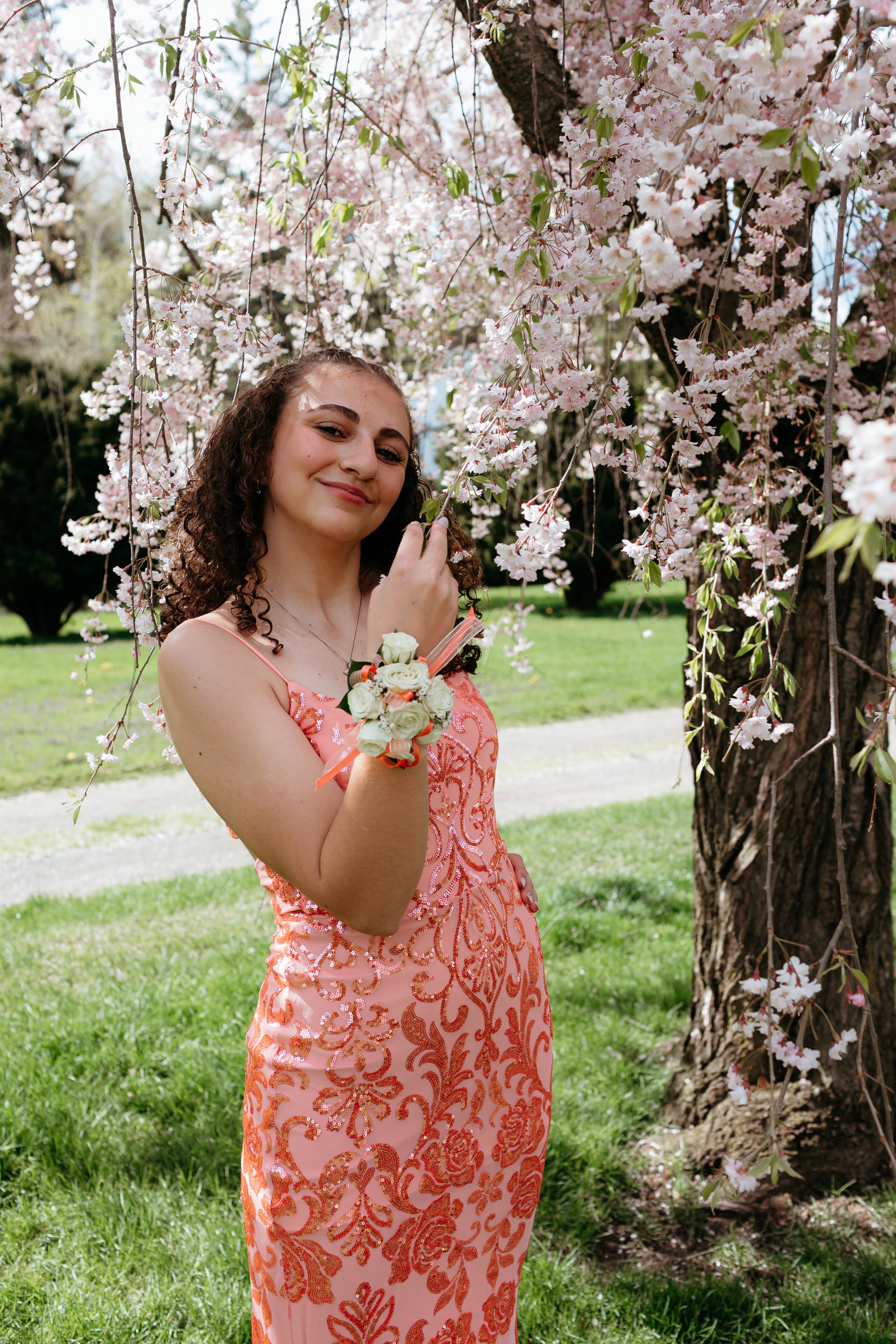 Young woman in an orange dress with floral patterns, wearing a corsage, standing under a blooming cherry blossom tree, smiling at the camera.