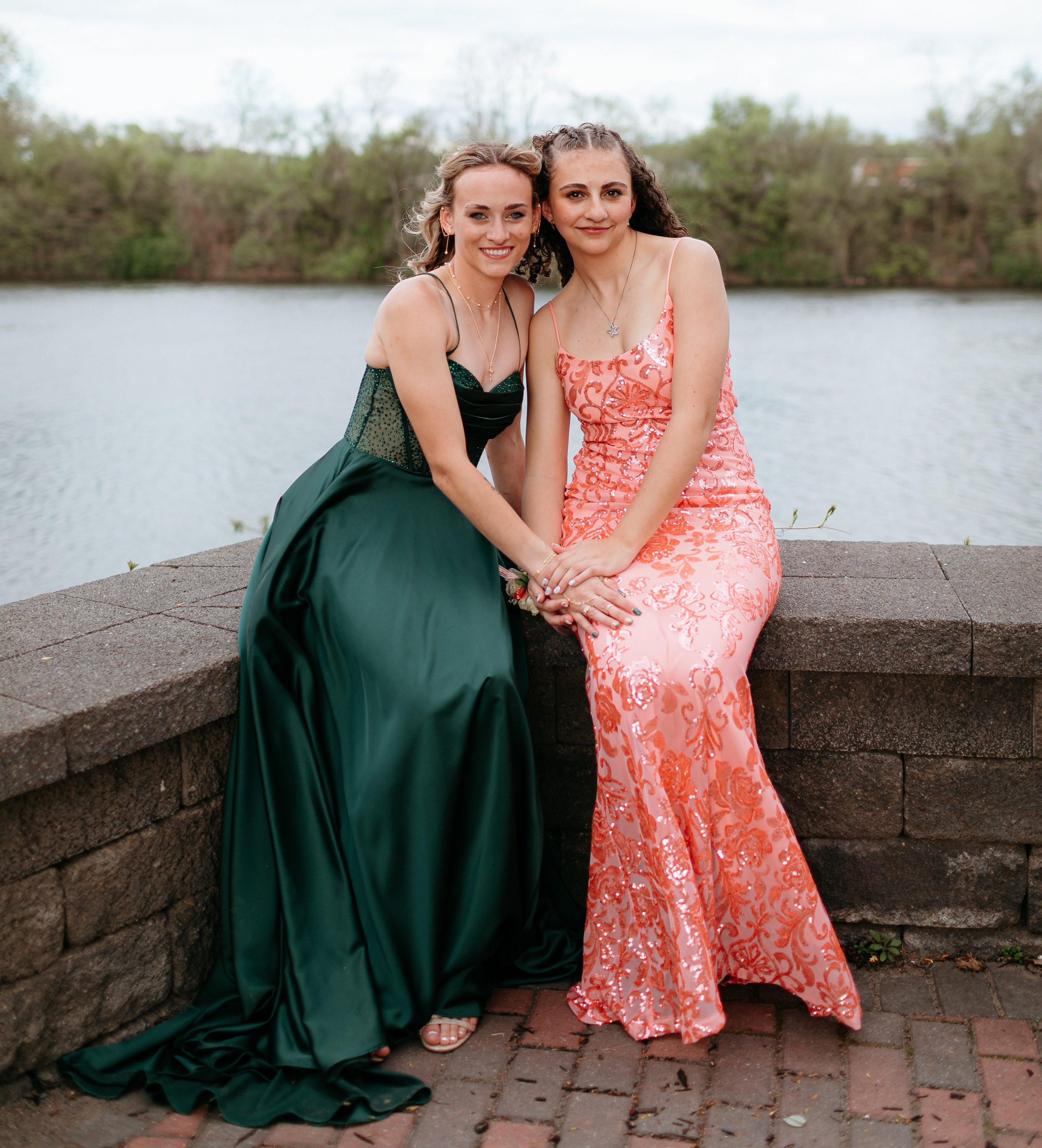 Two individuals in formal dresses sitting on a stone bench by a lake.