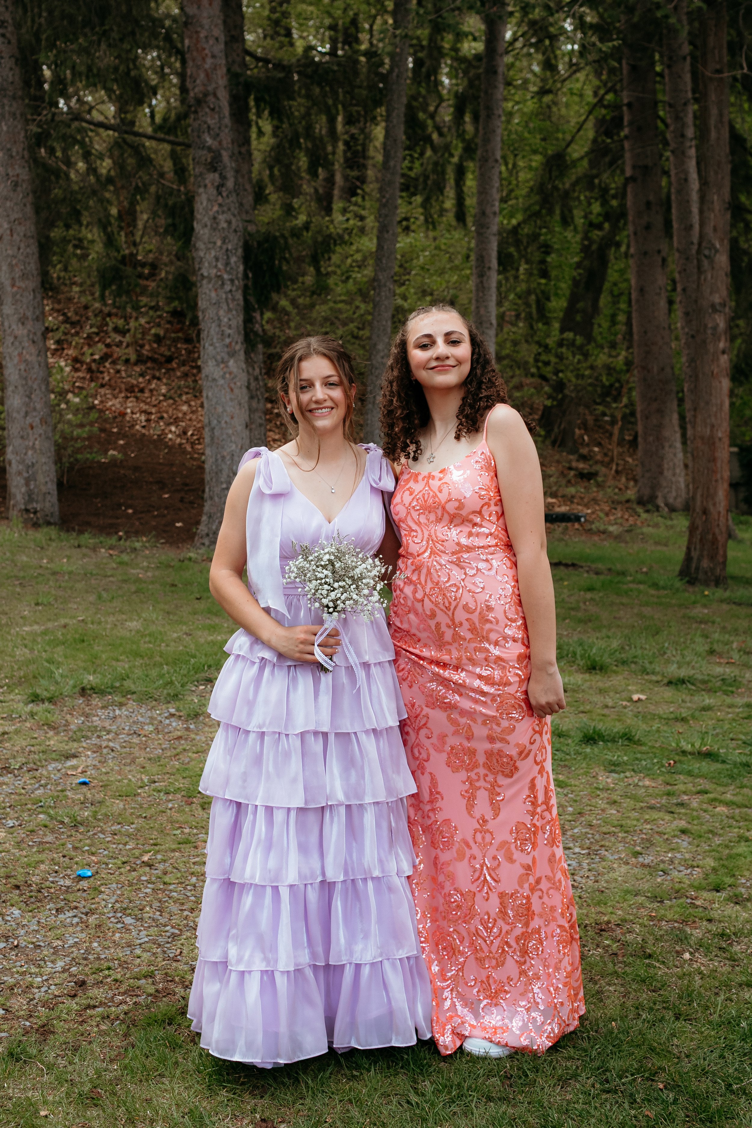 Two women in formal dresses pose outdoors, one in a lavender tiered gown holding flowers and the other in a pink and orange embellished gown, surrounded by trees.