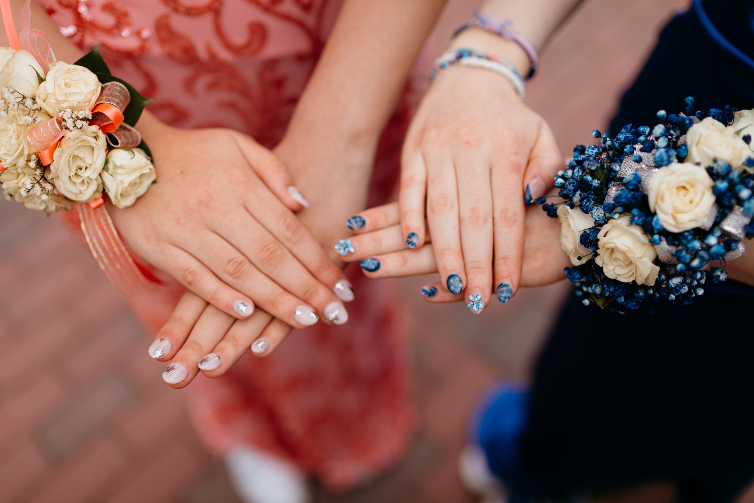 Close-up of two hands with decorated nails and corsages, one floral and blue, the other light with white and orange roses.