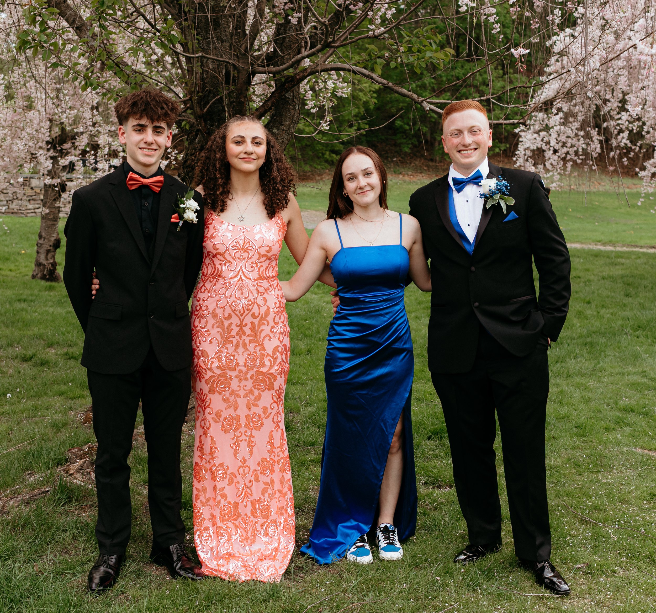 Four people in formal attire posing outdoors during spring, with trees and greenery in the background.