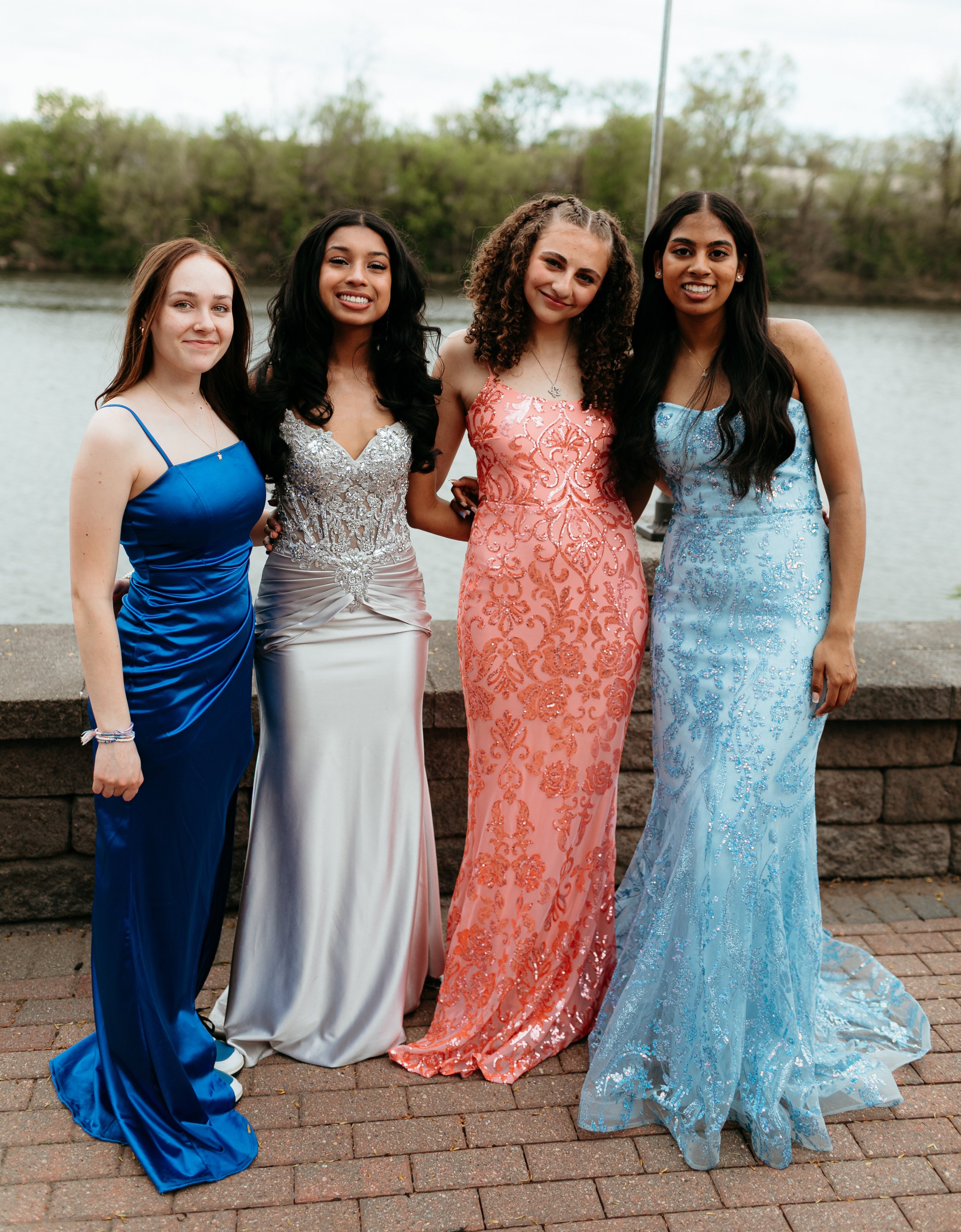 Four women in elegant evening gowns posing outdoors, with a background of a river and trees, wearing different colored dresses: blue, silver, pink, and light blue.