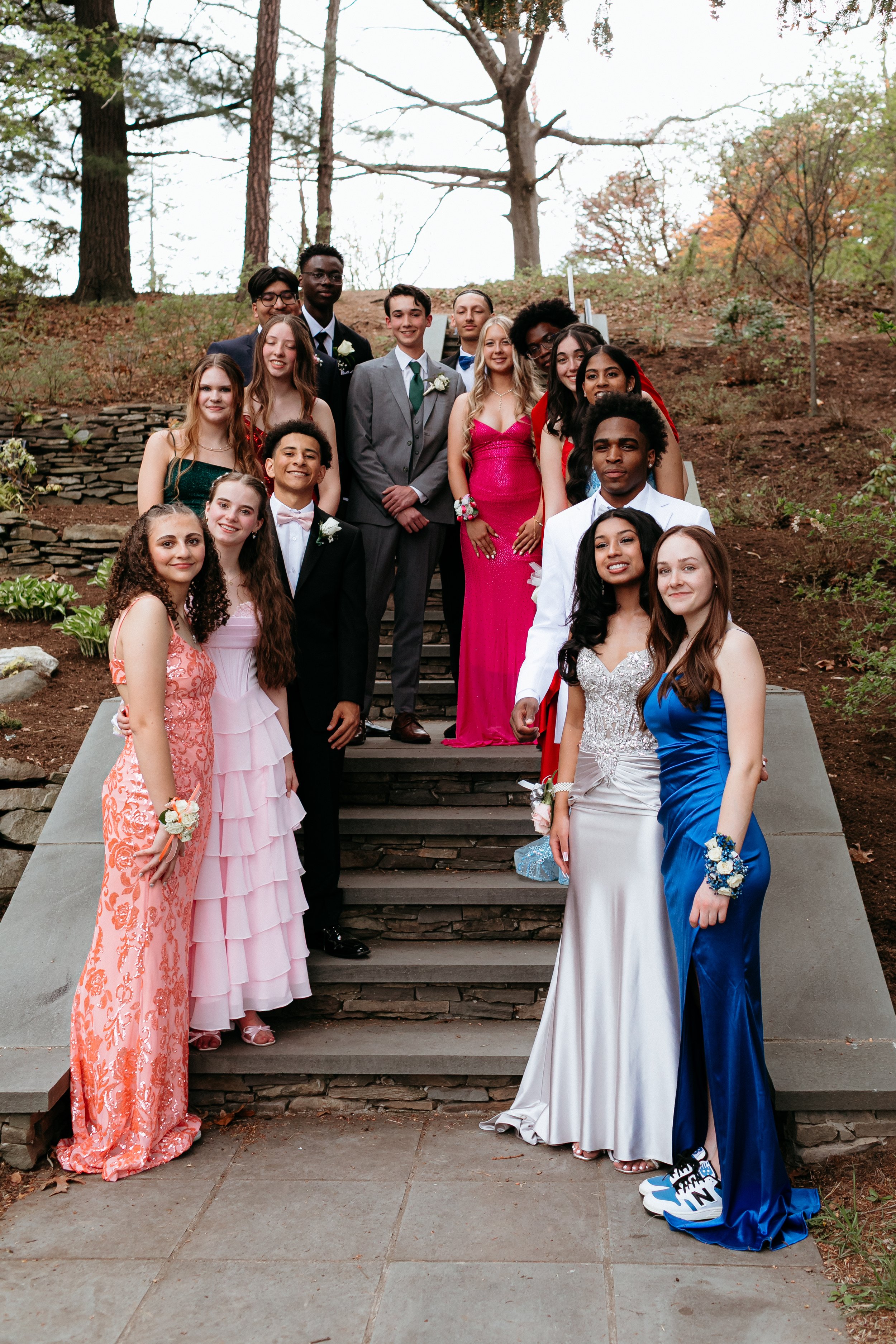 A group of teenagers dressed in formal attire, standing on outdoor steps, likely attending a prom or formal event. The young men are wearing suits, and the young women are in evening gowns with corsages. Trees and foliage are in the background.