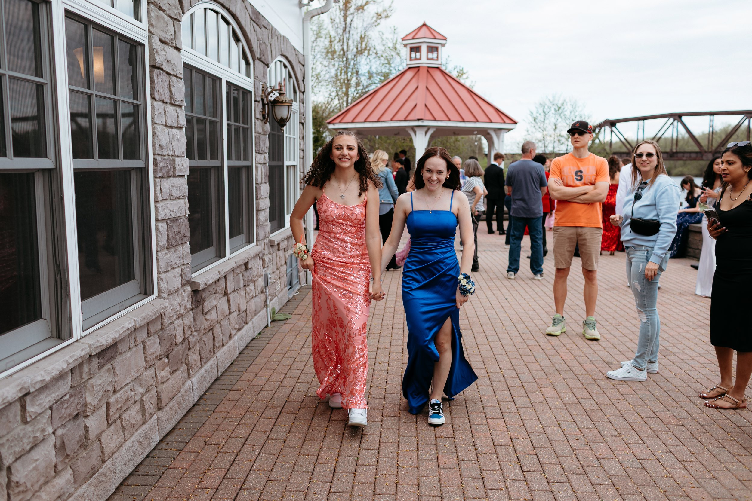 Two girls in formal dresses walking outside, one in pink and one in blue, surrounded by people in casual clothing, near a building with brick and stone elements.