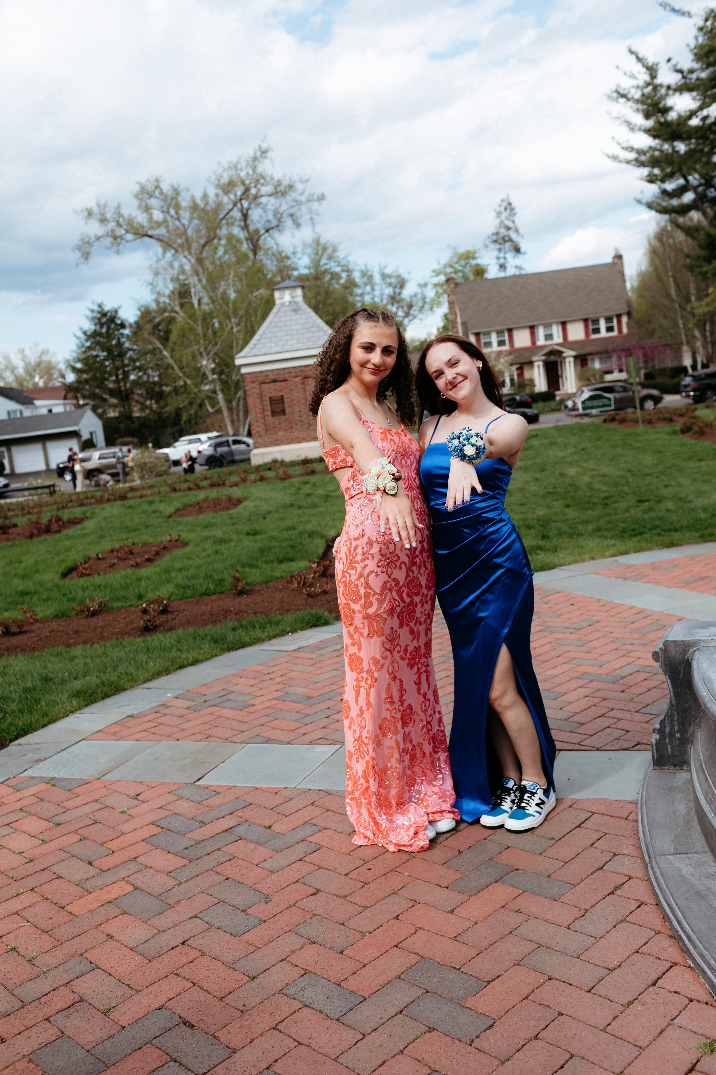 Two young women in formal dresses, one in orange with floral patterns and the other in royal blue, stand on a brick walkway. They both display their corsages and wear sneakers. The background features a grassy area, trees, and houses.
