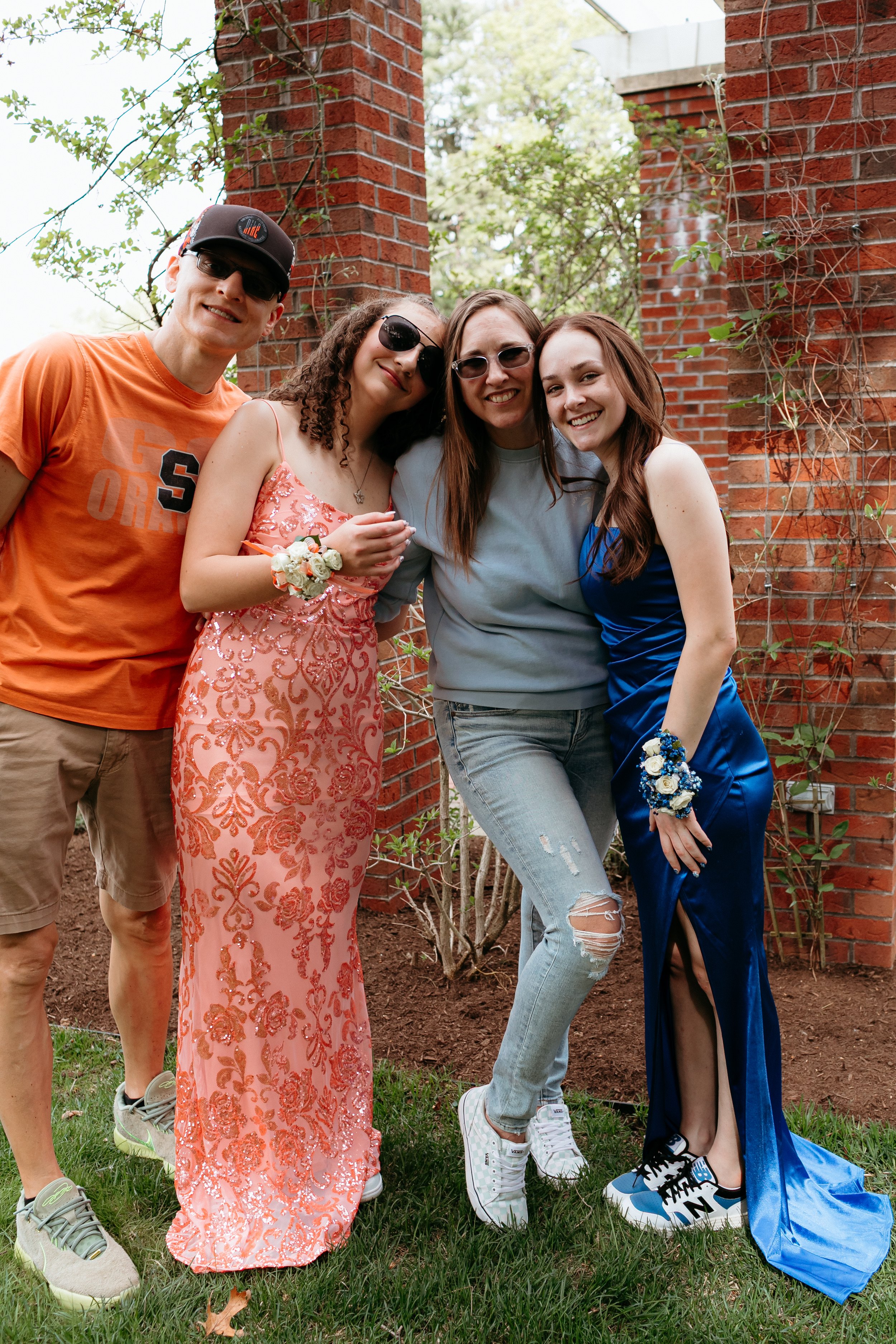 A group of four people posing outdoors by a brick column. Two individuals wear formal gowns with corsages: one in peach and the other in royal blue, while the other two are casually dressed. They all appear to be smiling and enjoying the moment.