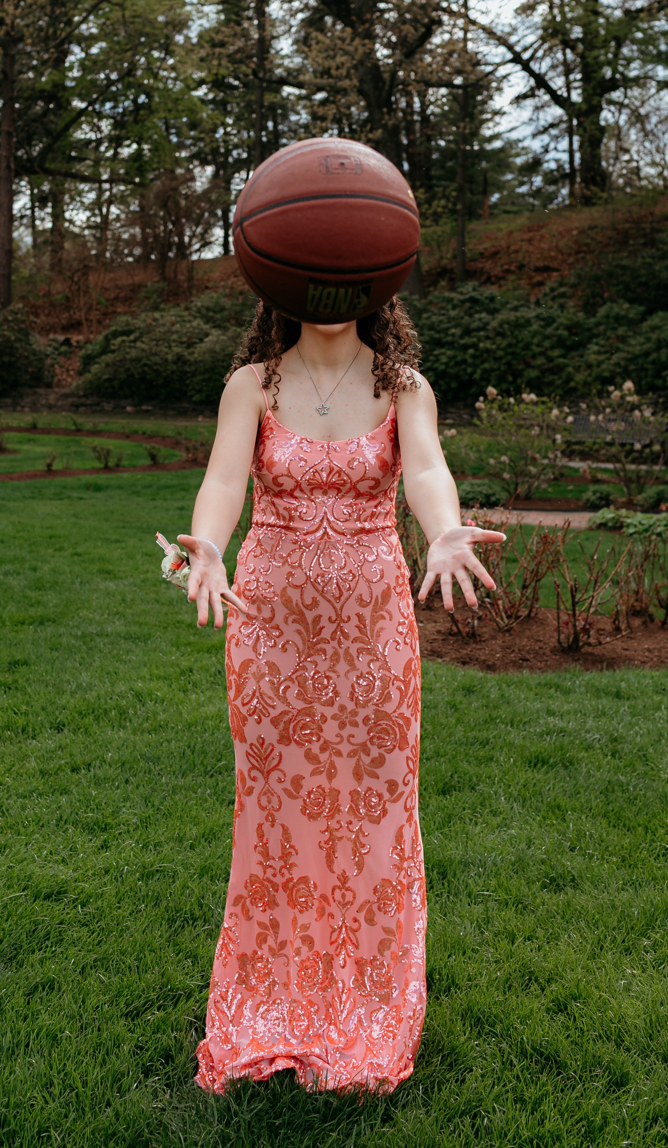 A person in a patterned coral dress is in a grassy area holding a basketball in front of their face.