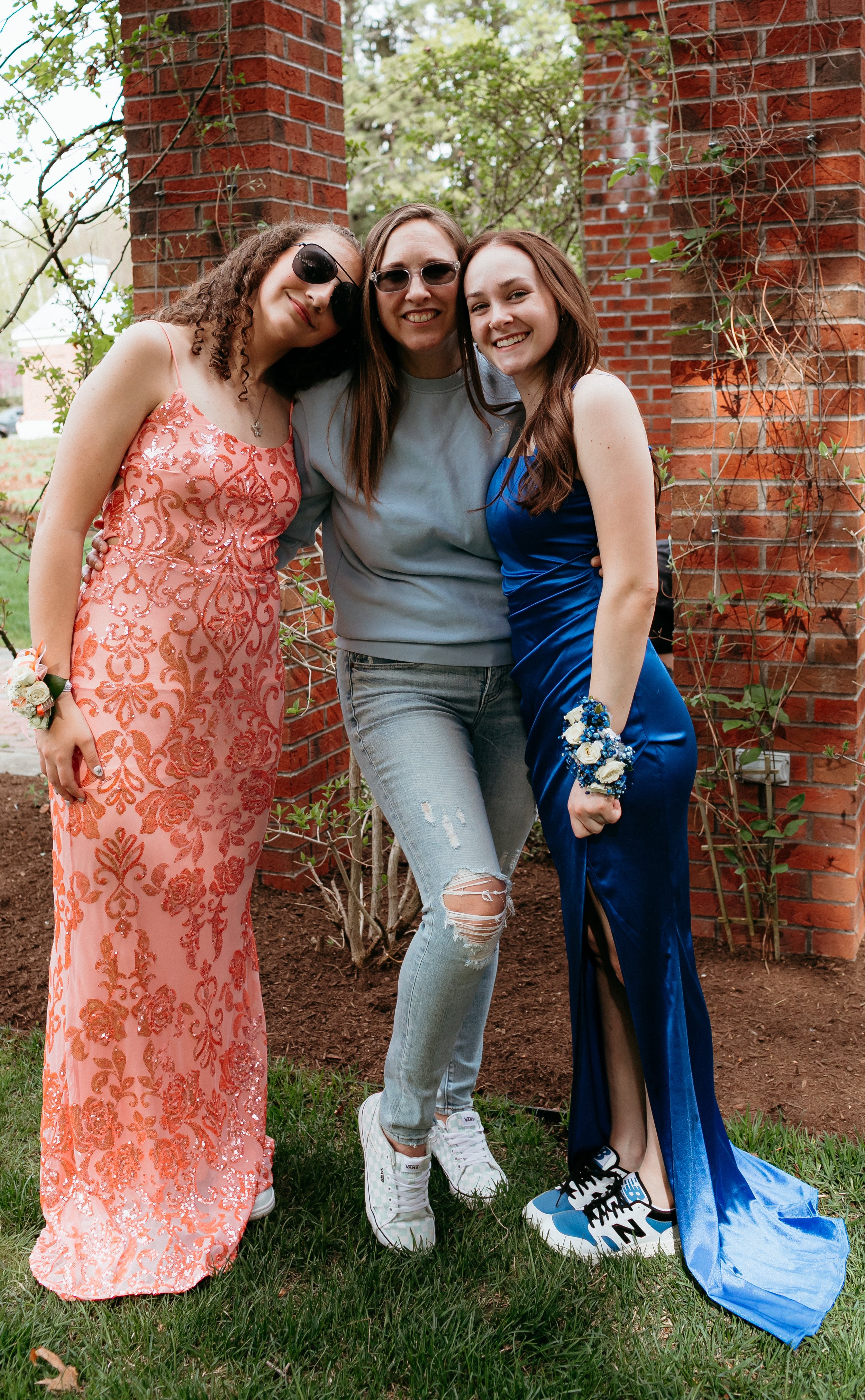 Three women posing for a photo; one in a pink dress, another in a blue velvet dress, and the third in casual wear with sunglasses, smiling against a brick and greenery background.