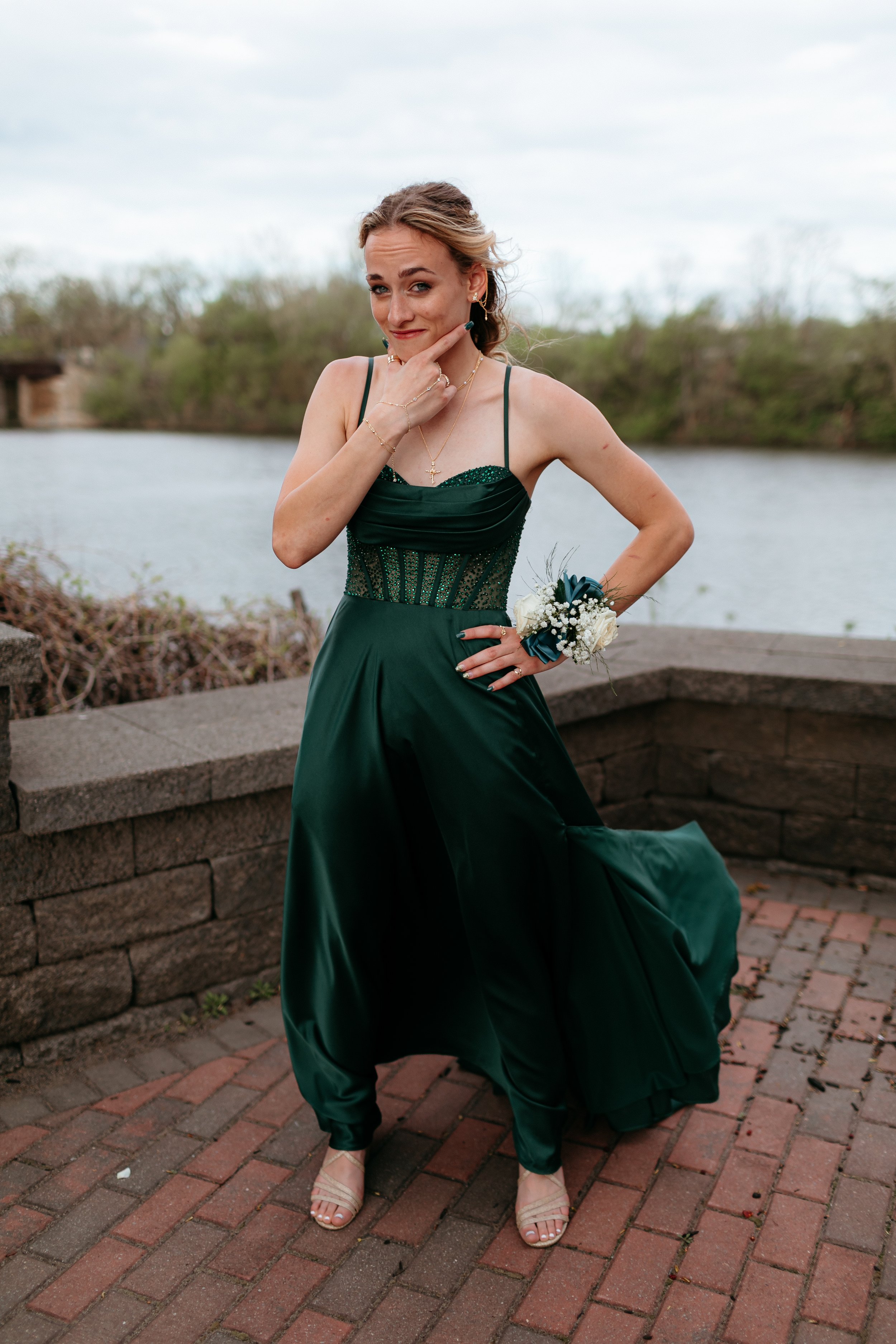 A woman in a dark green formal dress posing playfully, wearing a floral corsage on her wrist, standing on a brick pathway near a body of water and greenery.