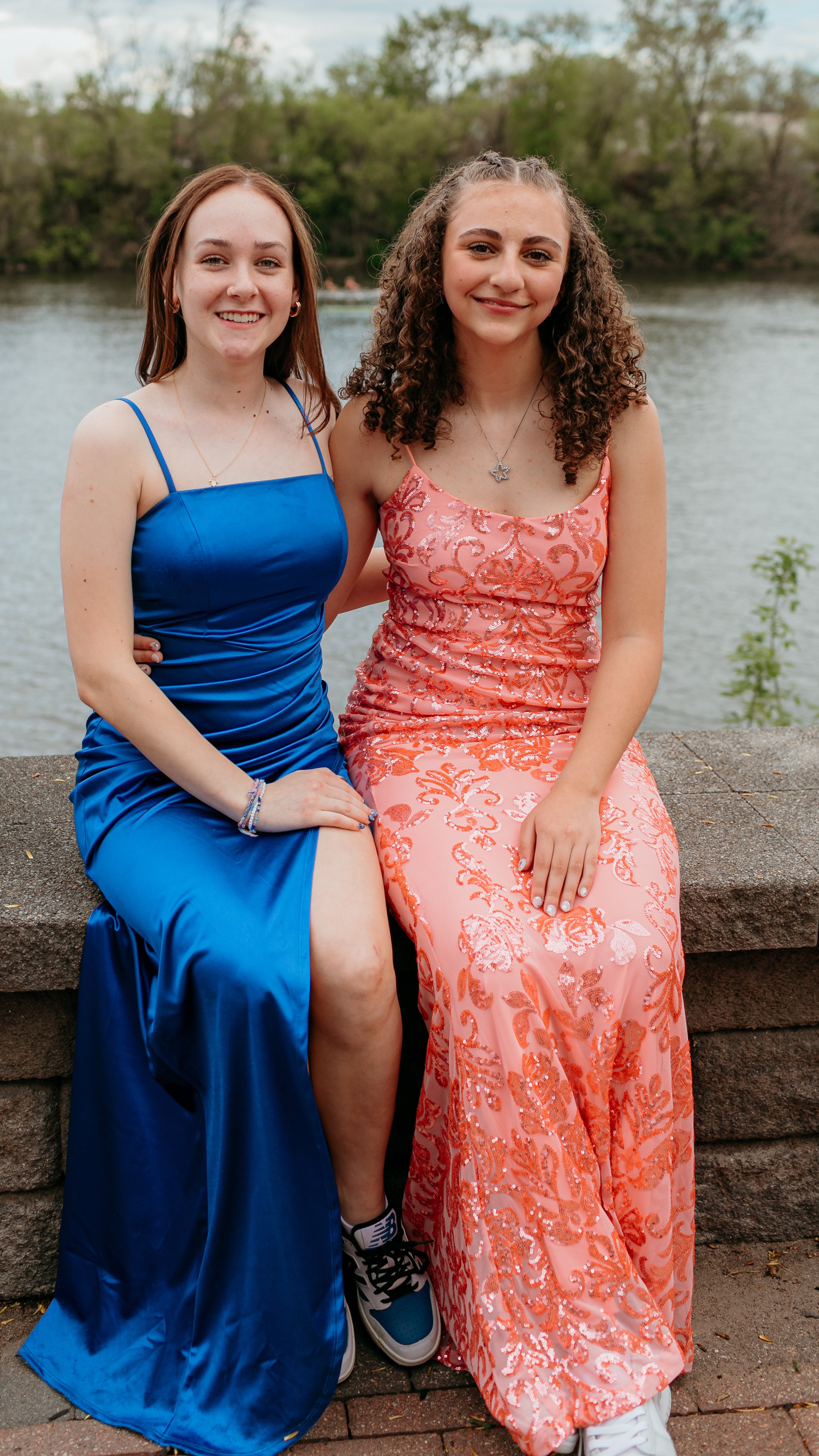 Two young women in elegant dresses sitting by a river, with trees and water in the background. One wears a blue dress and the other a pink dress with floral patterns.