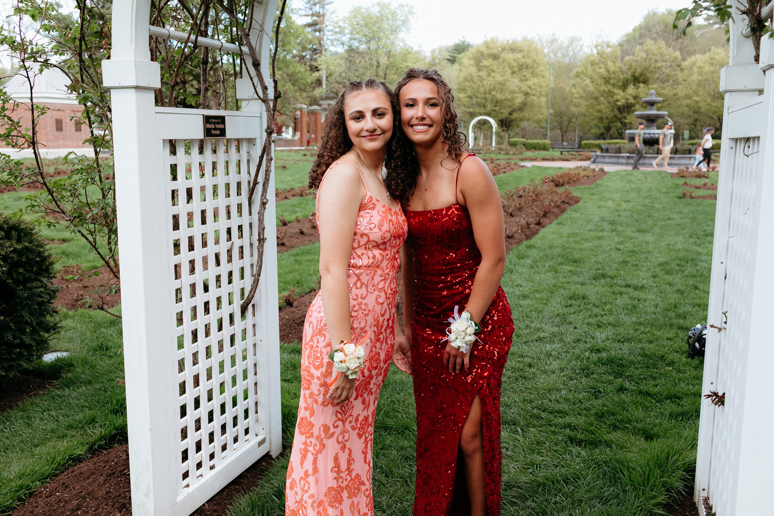 Two young women in formal dresses, one in orange and one in red, standing in a garden under a white archway. Both wear corsages.