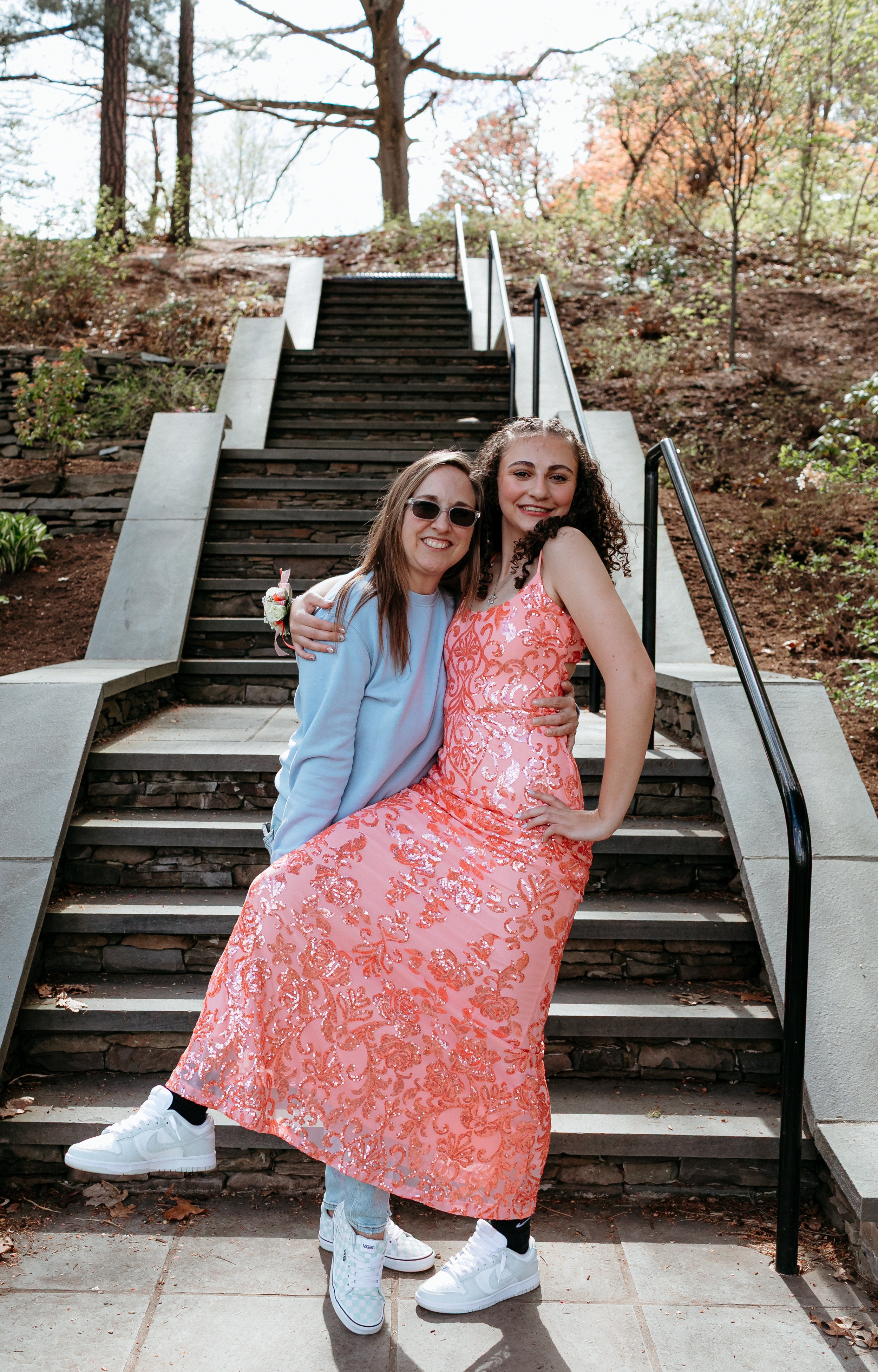 Two smiling individuals posing on outdoor steps, one wearing sunglasses and a blue top, the other in a pink floral dress with sneakers, surrounded by trees and greenery.