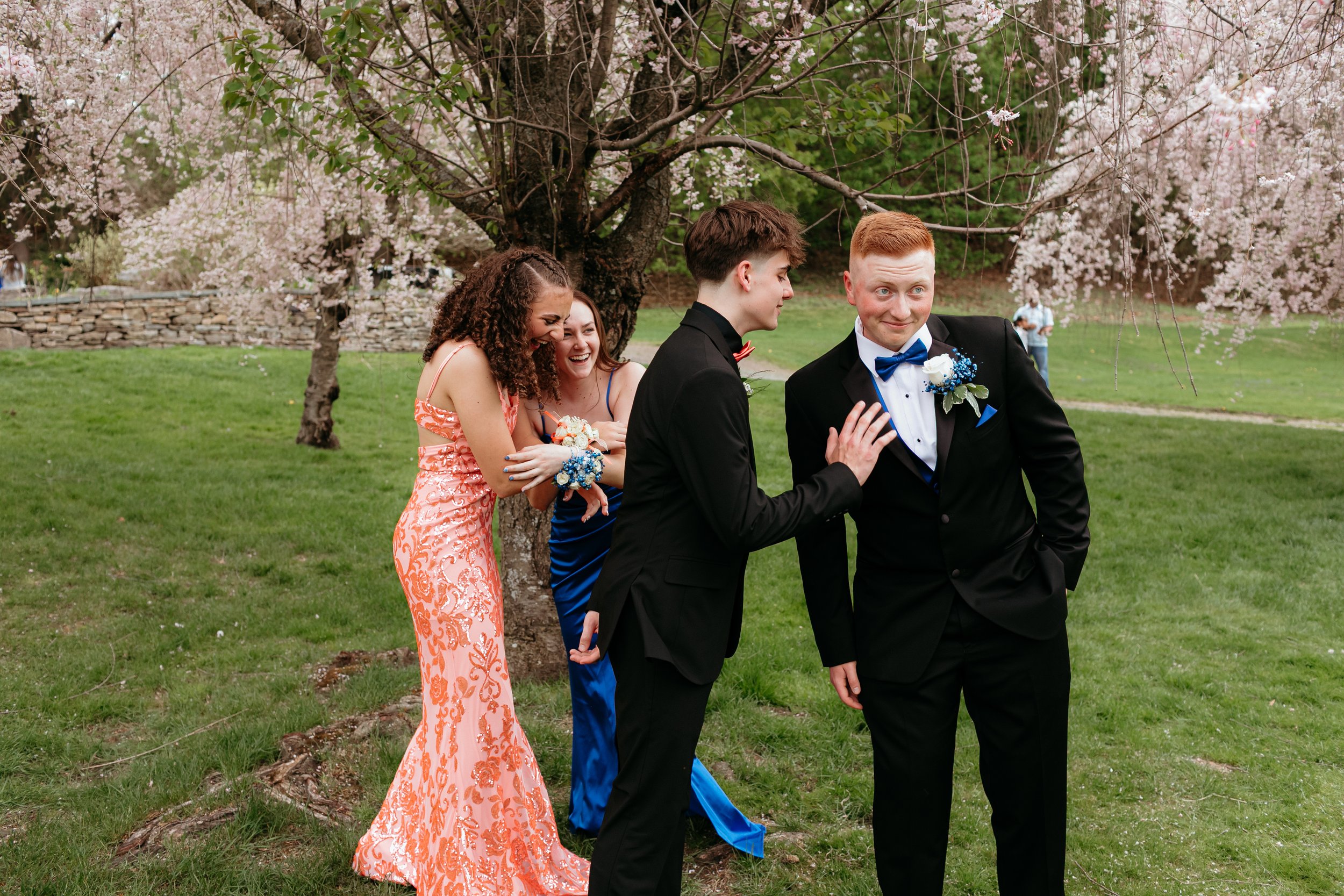 Prom photoshoot of group of friends in formal attire laughing in a park with cherry blossom trees