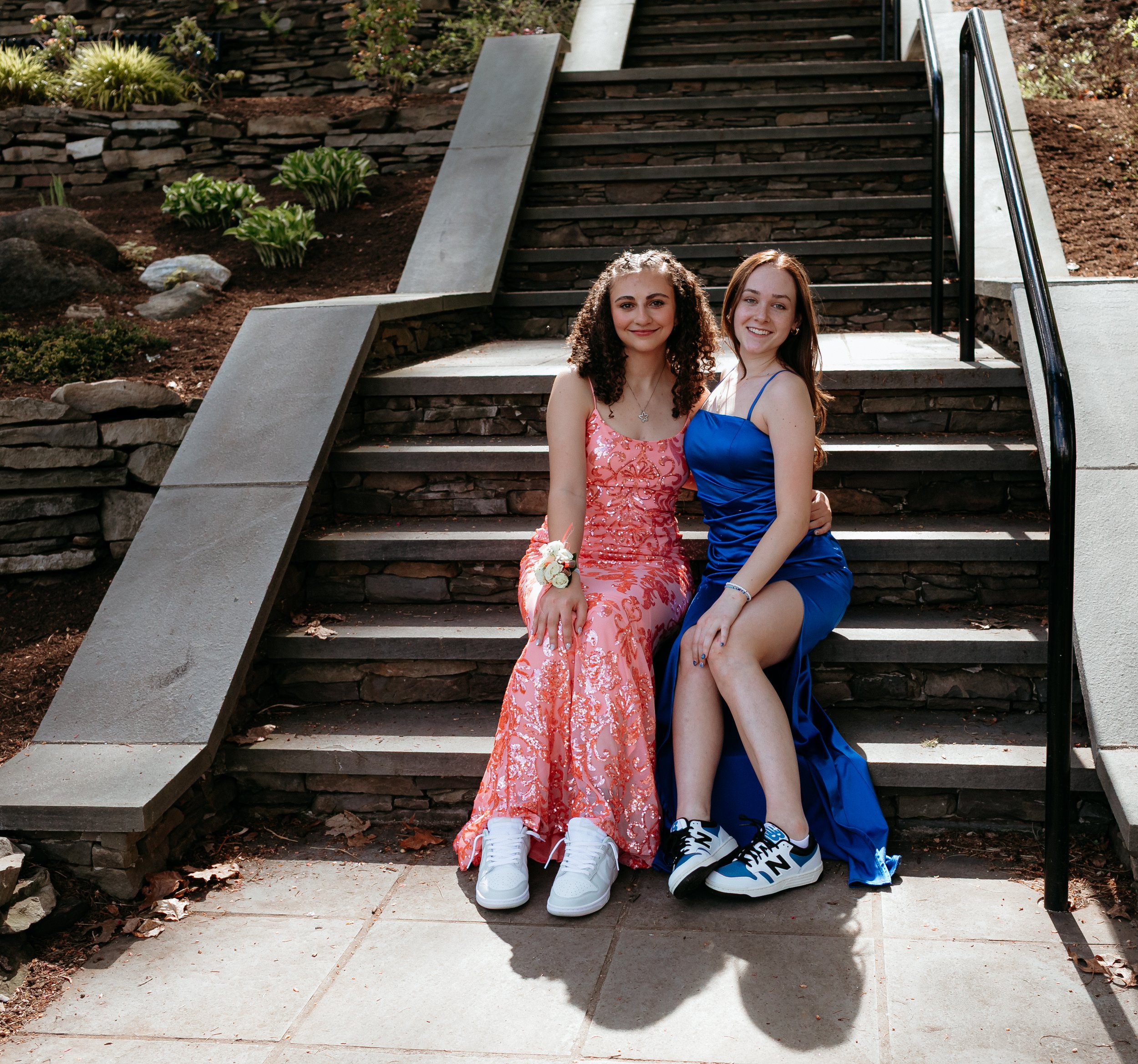 Two young women in formal dresses sitting on outdoor stairs, wearing sneakers, with a garden background.