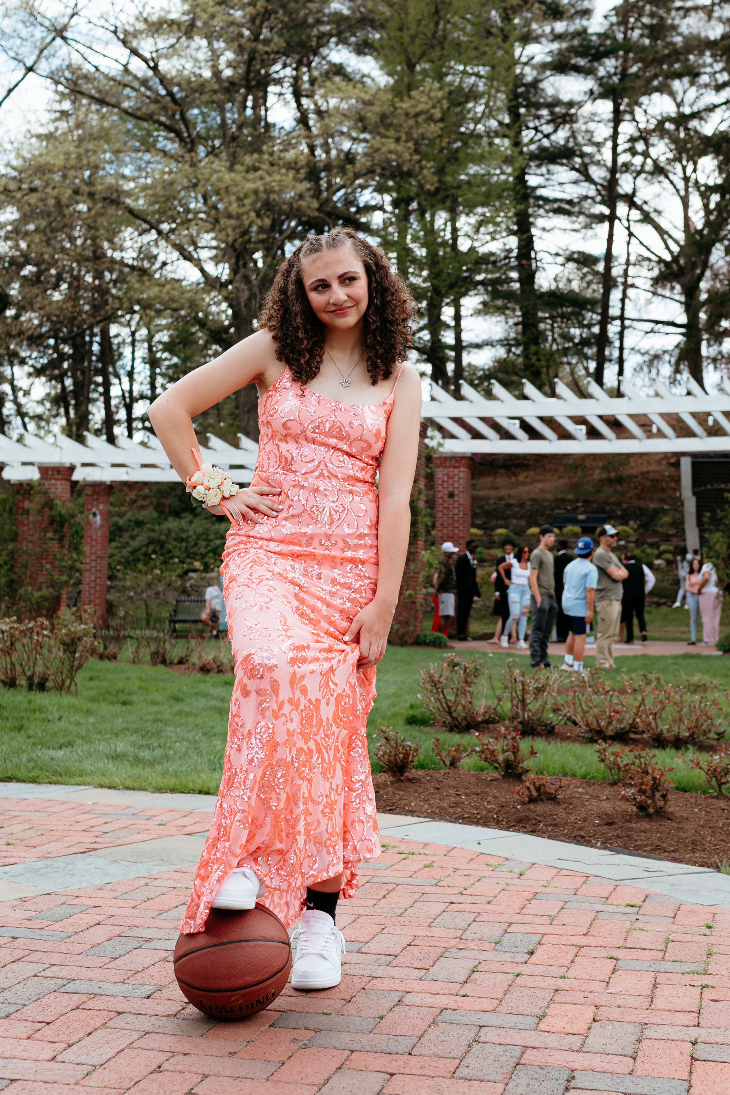 A young woman in a floral pink dress stands on a brick pathway with a basketball beneath her foot, surrounded by greenery and people in the background.