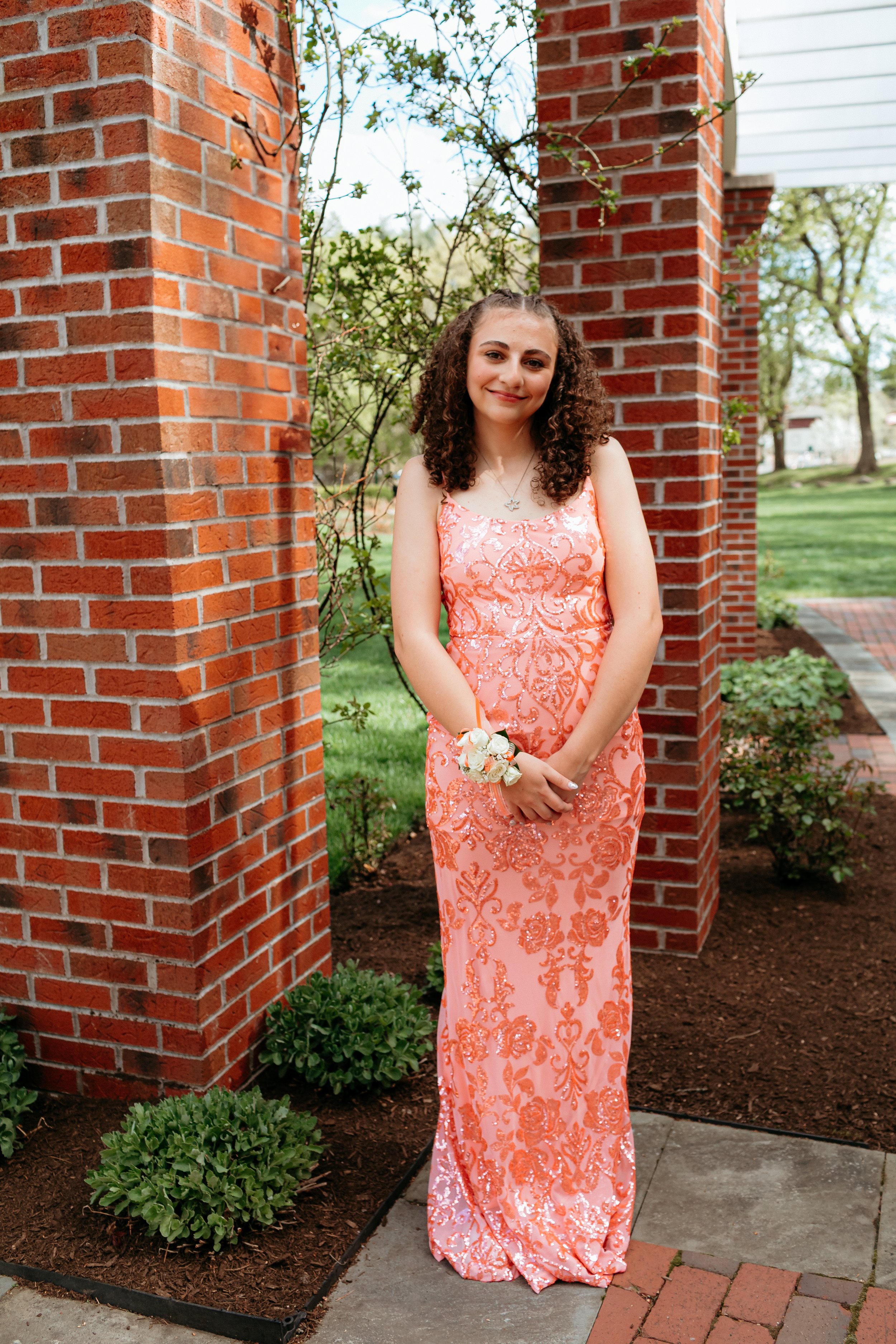 Young woman in a peach embellished gown standing by a brick pillar with greenery and garden background.